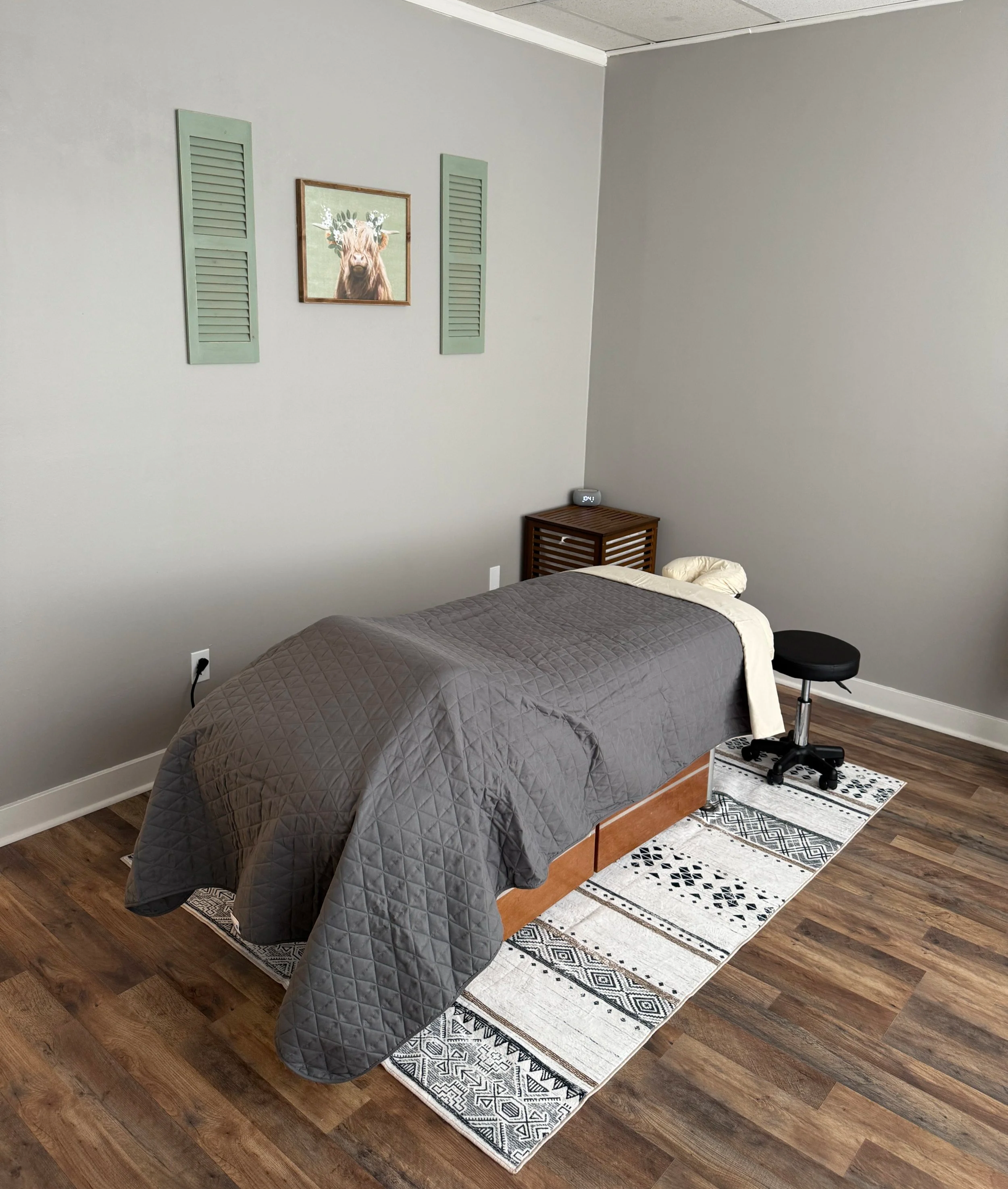 A photo of Rae Haven's Ellensburg massage studio, featuring a massage table with grey bedding, wall decor, a patterned rug, and essential oils.