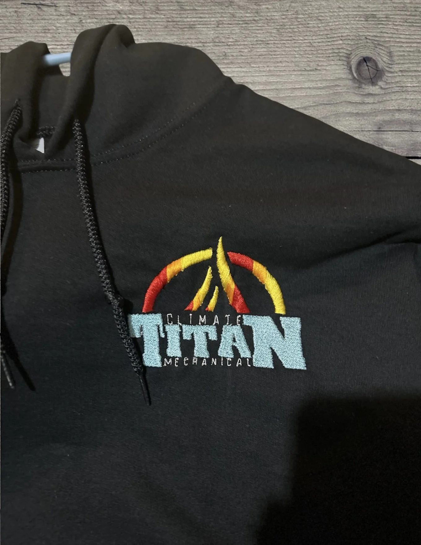 Close-up of a black hoodie with a colored embroidered logo that reads 'Climate Titan Mechanical' and a design of three yellow and red flame-like shapes.