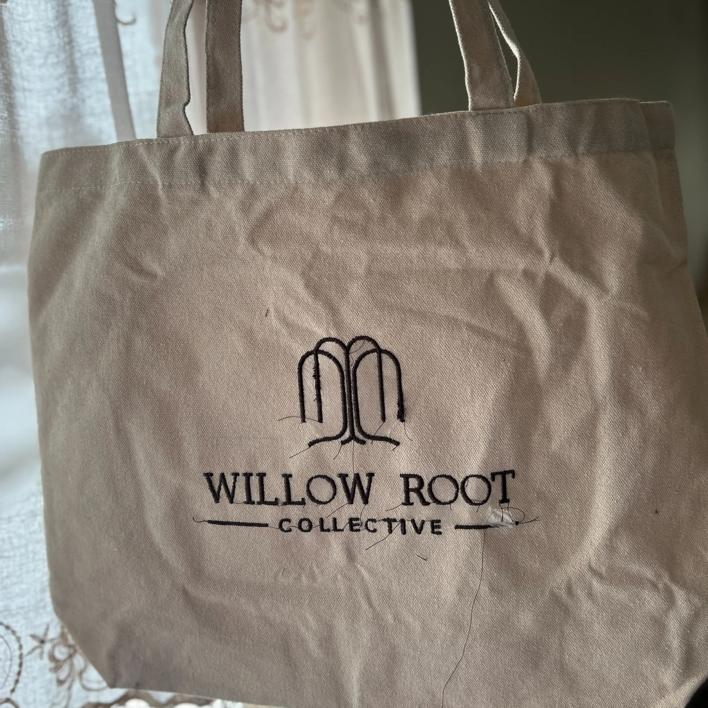 A beige tote bag with the Willow Root Collective logo embroidered in black, featuring hands in a prayer position above the text.