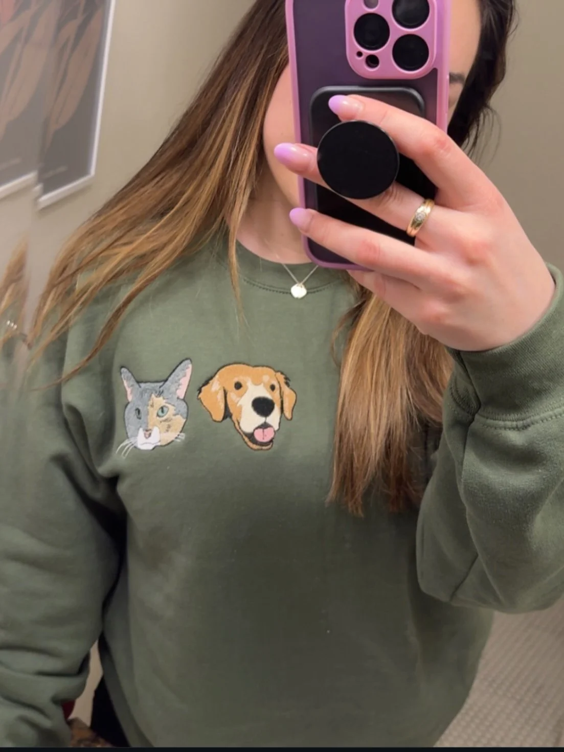 A woman taking a mirror selfie with a pink phone in a case with a black grip. She is wearing a green sweatshirt with embroidered patches of a cat and a dog on the chest area, a silver ring on her finger, and a small necklace. Her hair is long and reddish-brown.