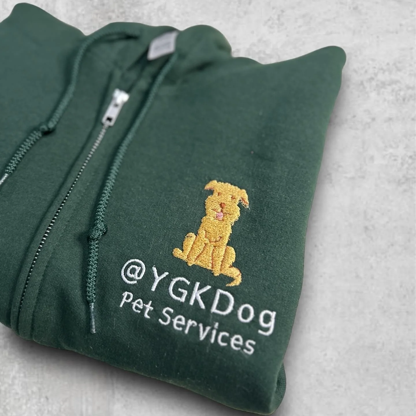 Green hoodie with embroidered logo of a yellow dog and text '@YGKDog Pet Services' on a light-colored surface.