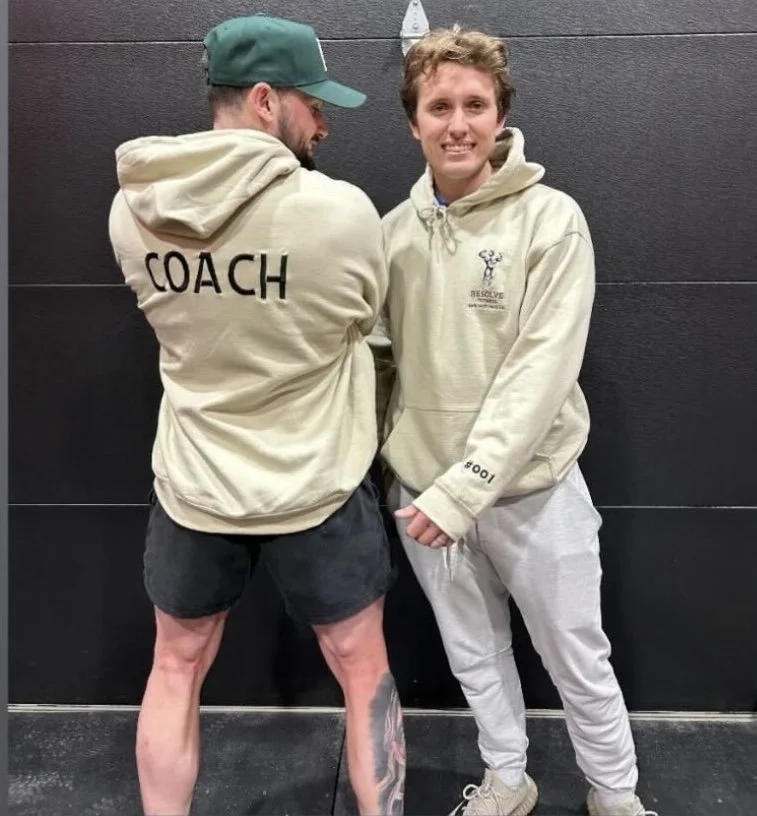 Two people standing against a dark wall. The person on the left has a beard, is wearing a green cap, beige hoodie with the word 'COACH' on the back, and black shorts. The person on the right has short, curly hair, is smiling, and is wearing a beige hoodie and gray sweatpants.