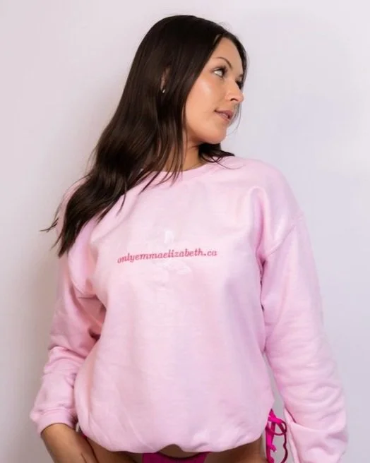 Woman with long dark hair wearing a pink sweatshirt with website text on it, standing against a plain white background.