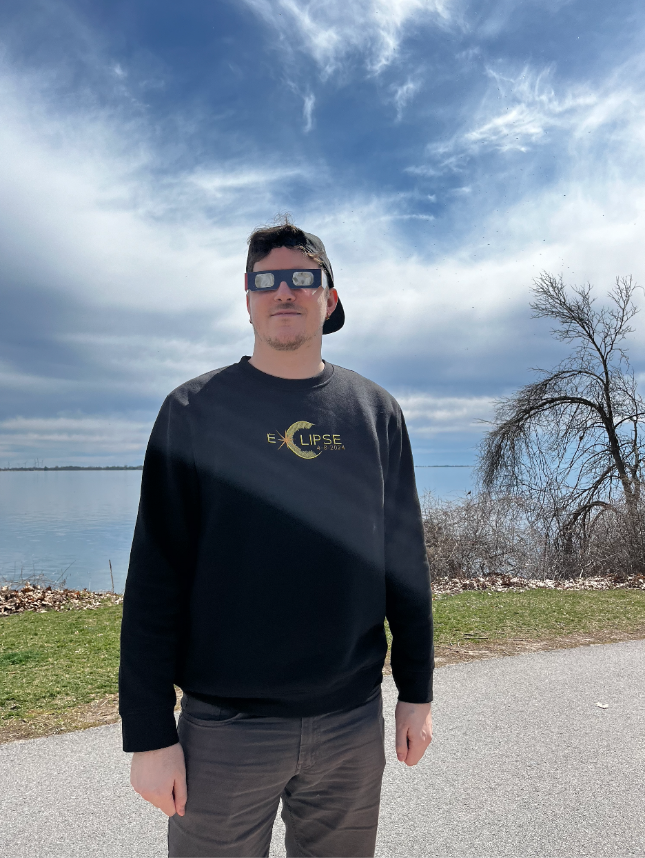 A man stands outdoors by a body of water, wearing eclipse glasses and a black sweatshirt with a lunar eclipse graphic and text. The sky is partly cloudy with the sun shining, and there are leafless trees nearby.