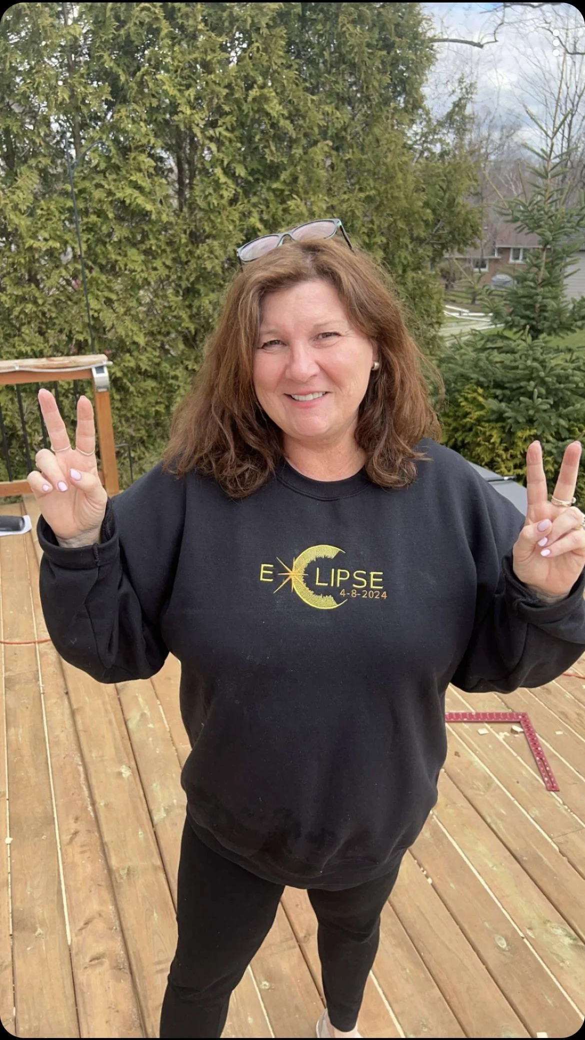 A woman is standing on a wooden deck outdoors, smiling and making peace signs with both hands. She has shoulder-length brown hair, is wearing glasses on her head, a black hoodie with a graphic that reads "ECLIPSE 4-8-2024" with a crescent moon, and b