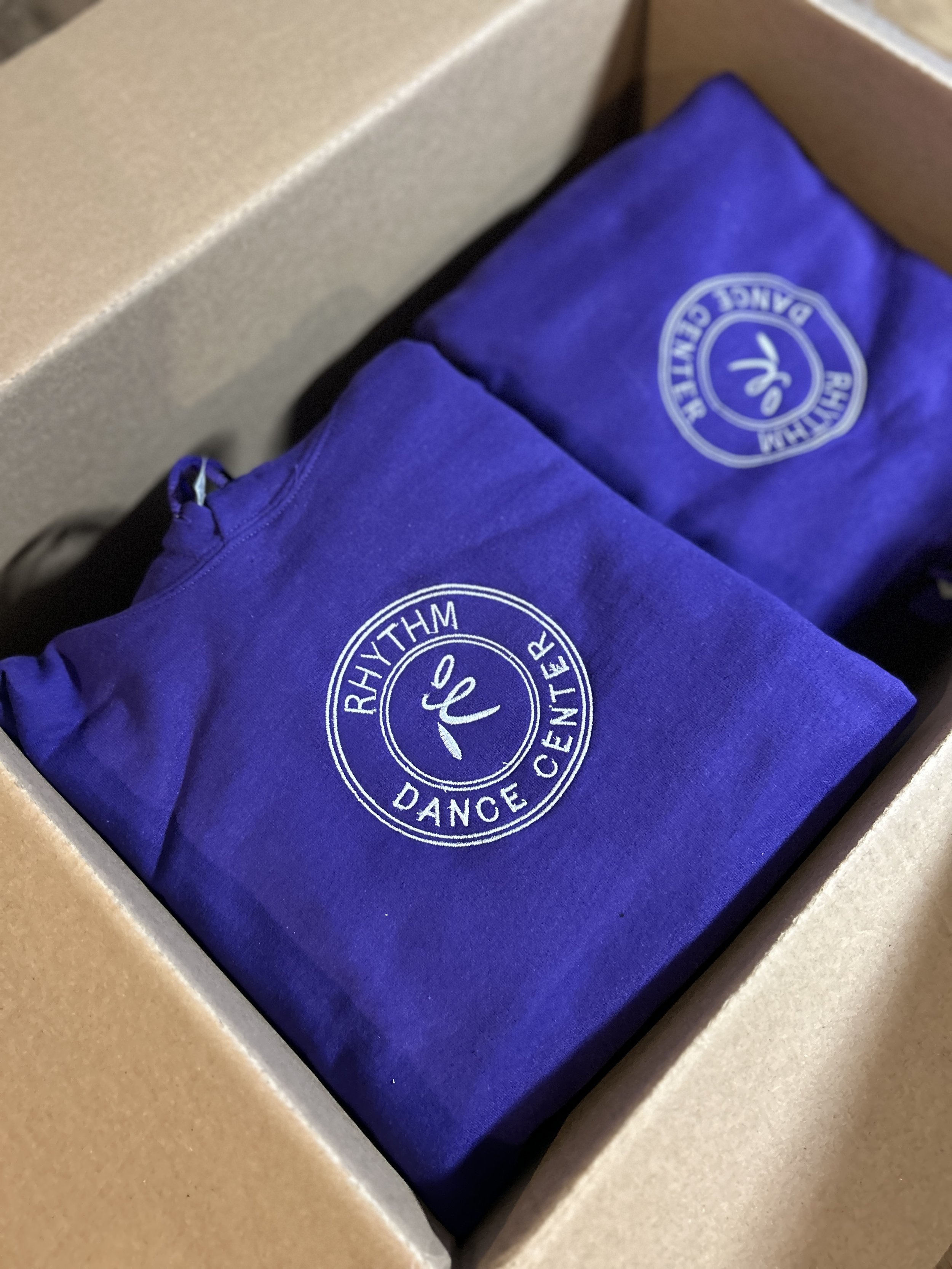 Two blue fabric items with 'Rhythm Dance Center' logo on them inside a cardboard box.