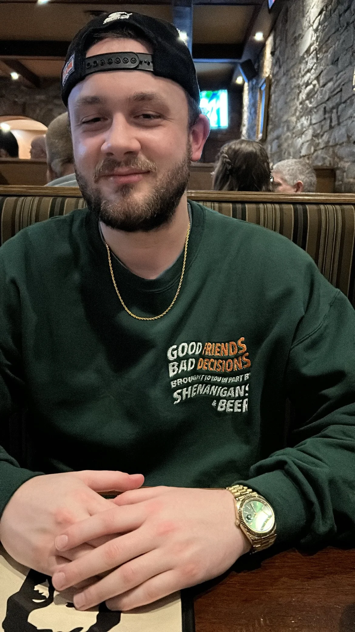 A young man with a beard, wearing a backwards cap, gold chain, and a green sweatshirt sitting at a restaurant table with a brick wall background.