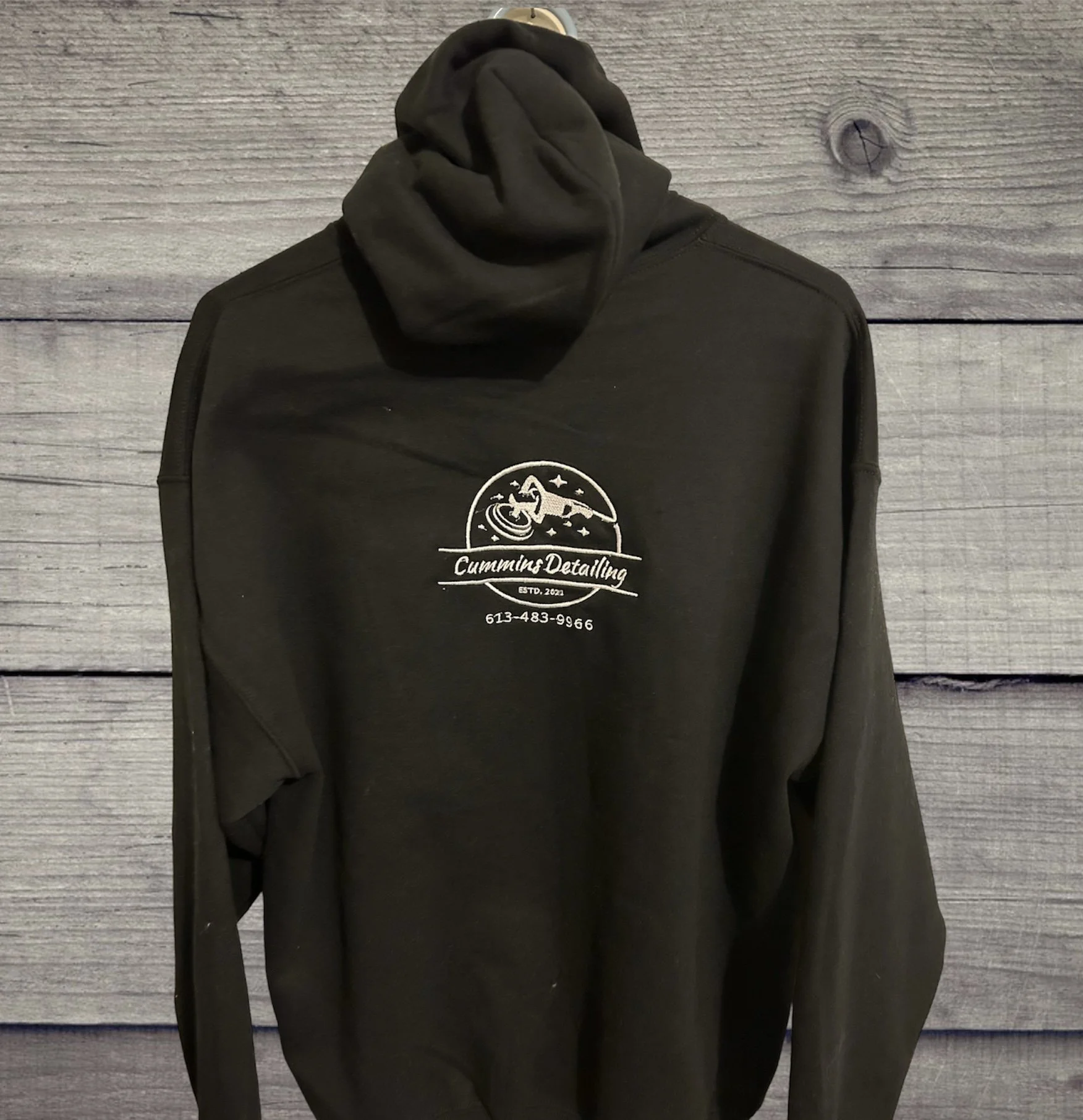 Black hoodie with logo and contact information hanging on a wall with wooden panel background.