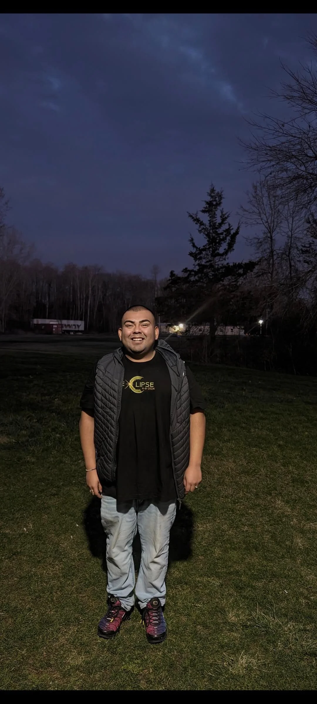 A young man smiling outdoors at night, standing on grass, with trees and a building in the background, under a dark sky.