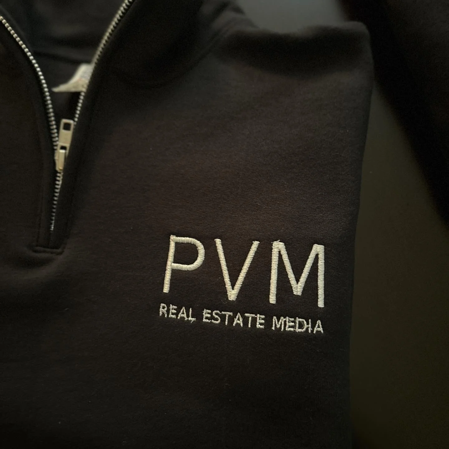 Close-up of a black jacket with a zipper and embroidered logo reading 'PVM' and 'Real Estate Media'.