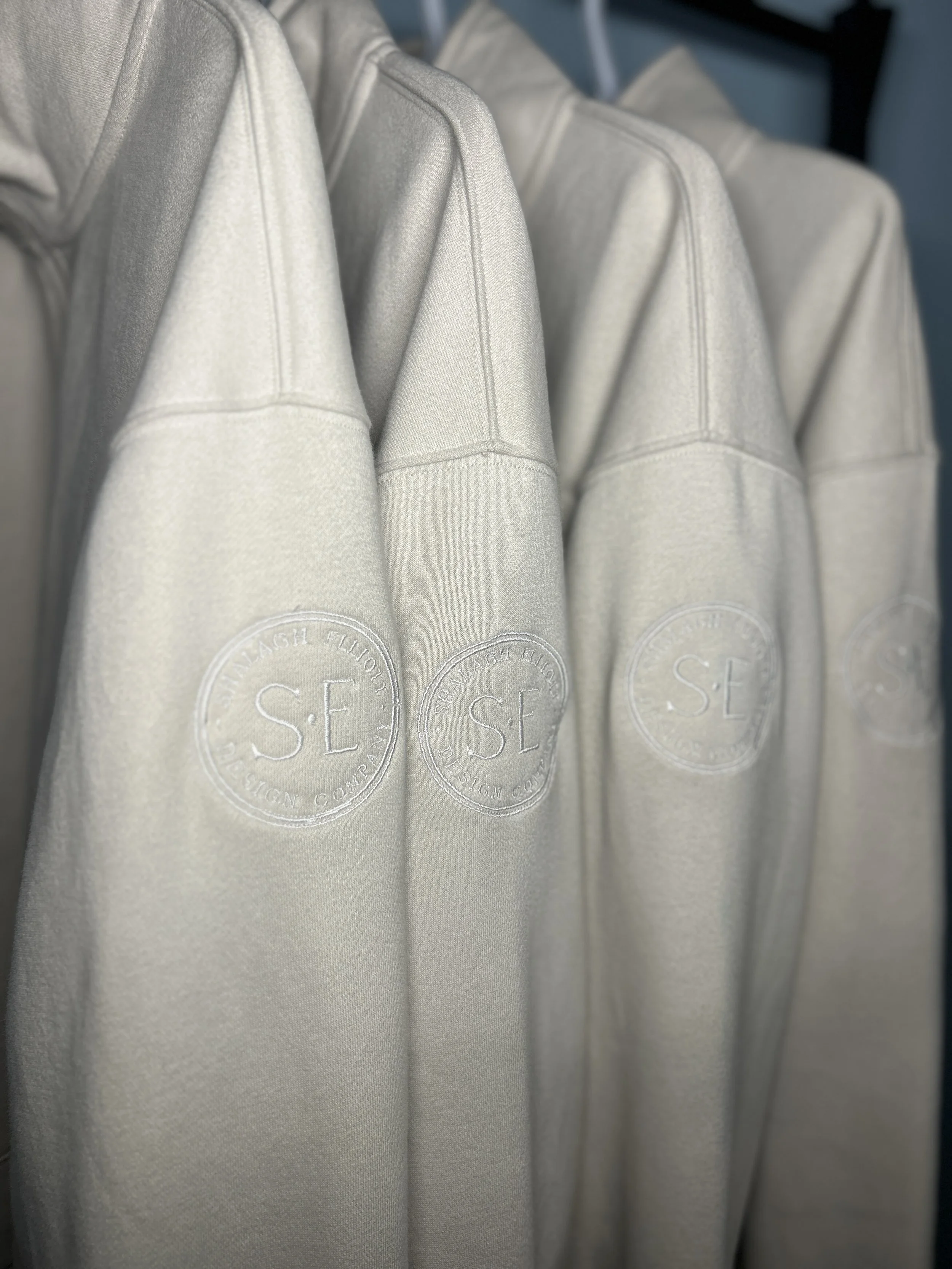 Four beige sweatshirts hanging on a rack, each with a circular patch embroidered on the chest with the initials "S E" and the text "STAY HIGHER THINK DESIGN COURT" around the circle.