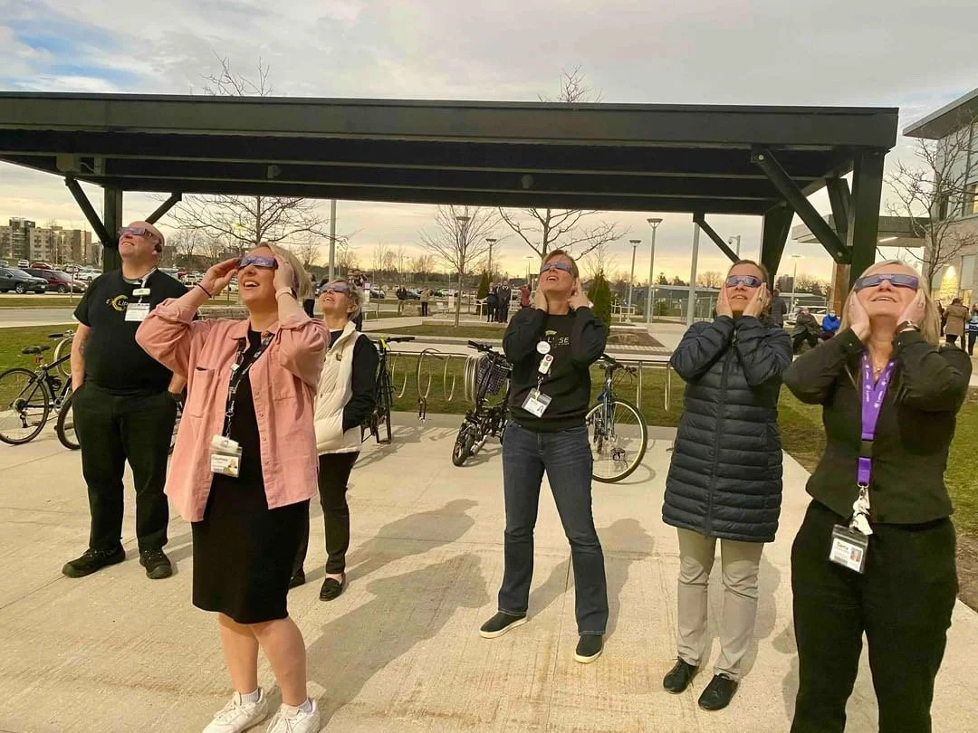A group of six adults wearing glasses and badges, standing outdoors and looking upward during daytime, with a bicycle rack and building in the background.