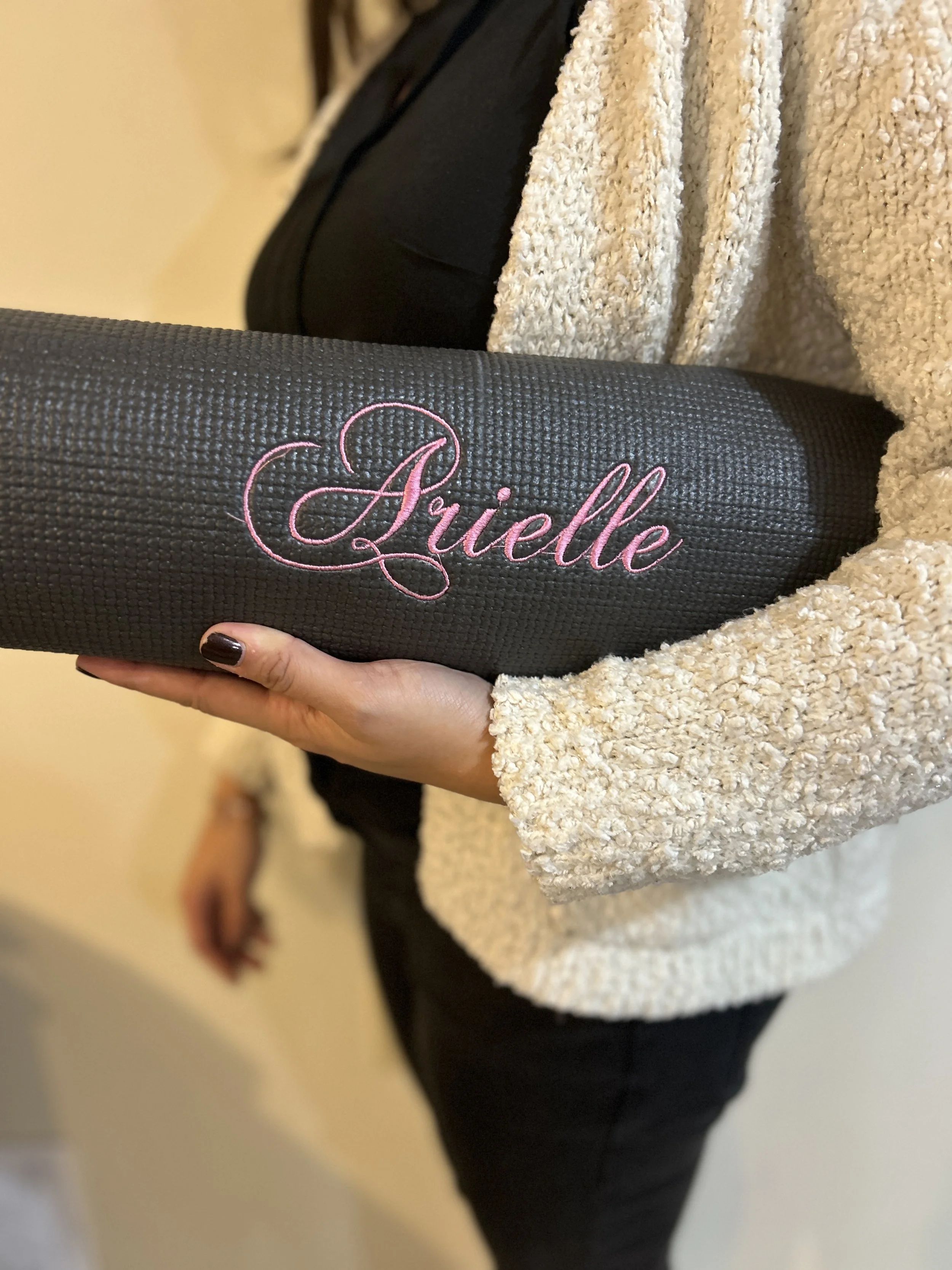 Person holding a black yoga mat with pink embroidered name 'Julie' in cursive, wearing a cream-colored sweater and black pants.