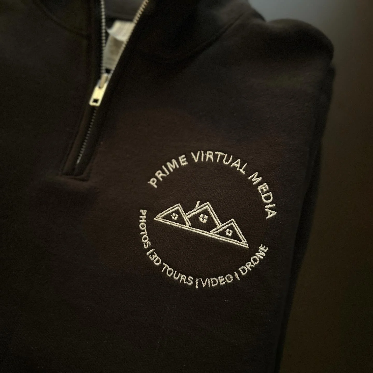 Close-up of a black fleece jacket with a zippered pocket featuring white embroidered text that reads "Prime Virtual Media" and "Get Yours Video | Drone" accompanied by a mountain range graphic.