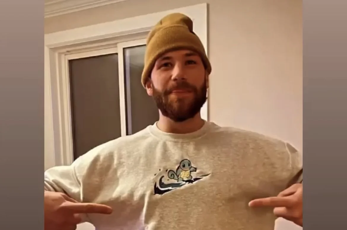 A man with a beard wearing a beige beanie and a gray sweatshirt featuring a cartoon squirrel on a tree branch points to his chest with both hands, standing inside a room near a window.