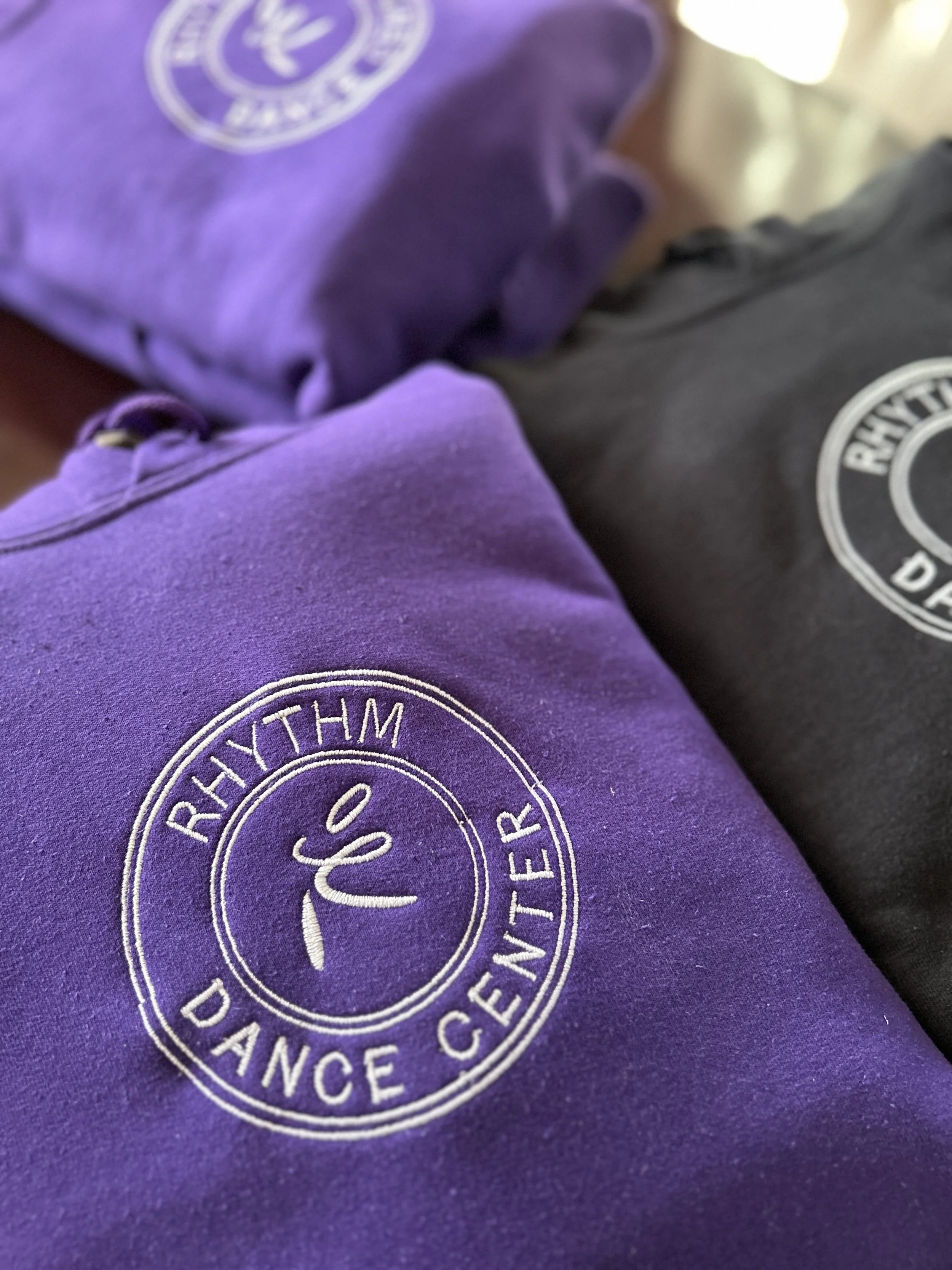 Close-up of purple and black sweatshirts with 'RhythM Dance Center' embroidered logo.
