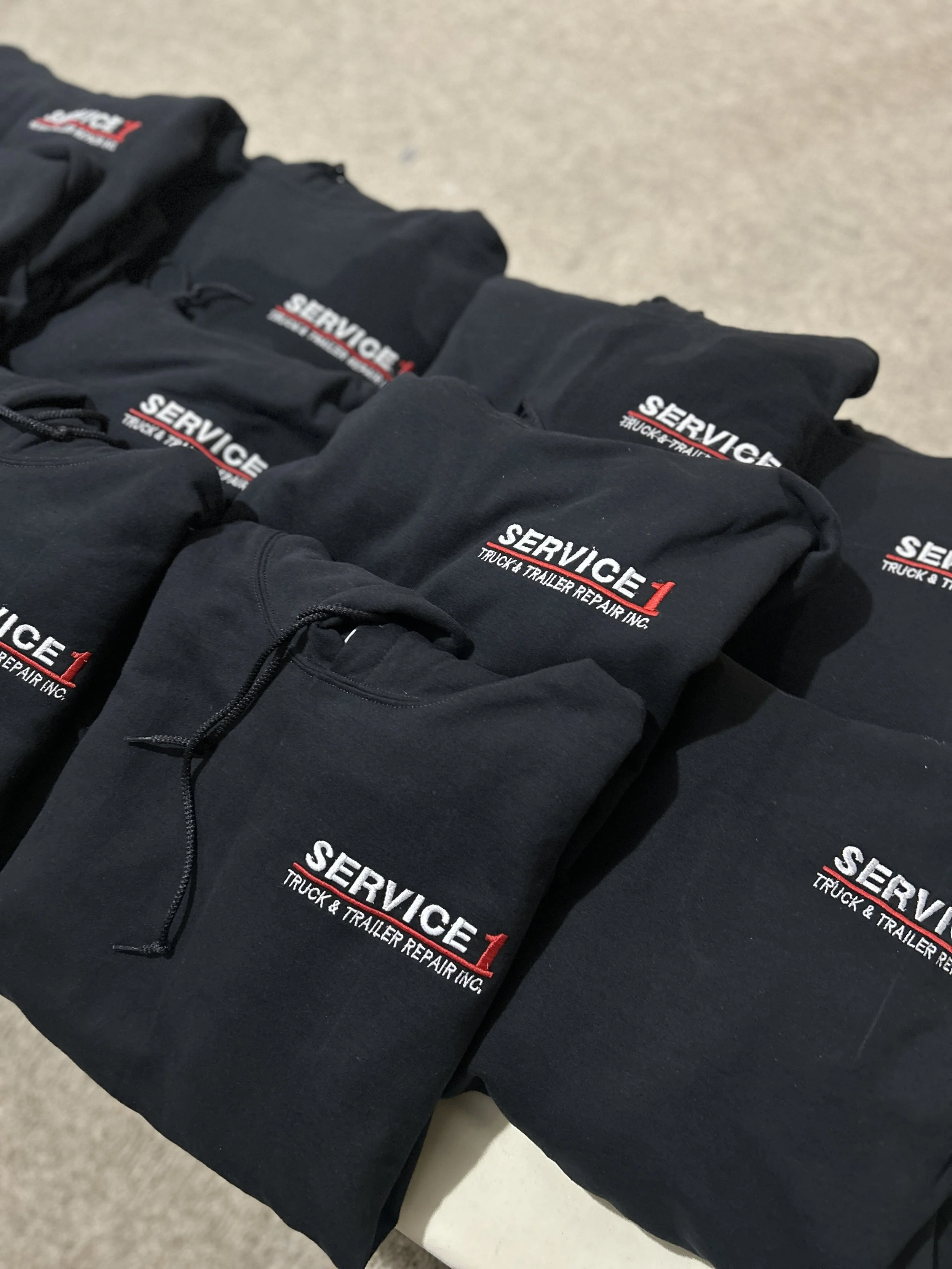 Black hoodies with embroidered logo of Service 1 Truck & Trailer Repair Inc., folded and arranged on a table.