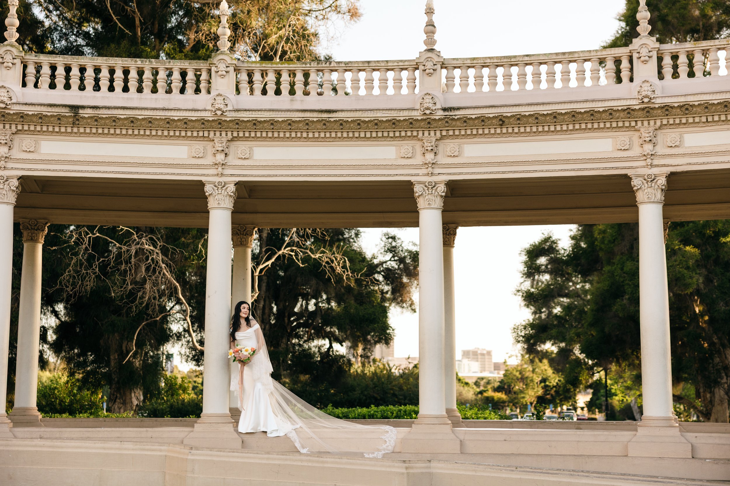 Bride in a long white dress with a veil holding a bouquet, standing in a classic architectural structure with columns and ornate carvings.