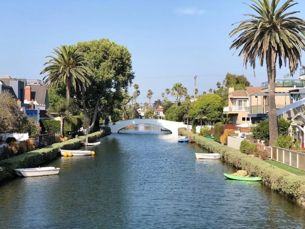 Scenic canal with boats, lush trees, and residential houses with balconies and gardens, under a clear blue sky, with a small white bridge in the background.