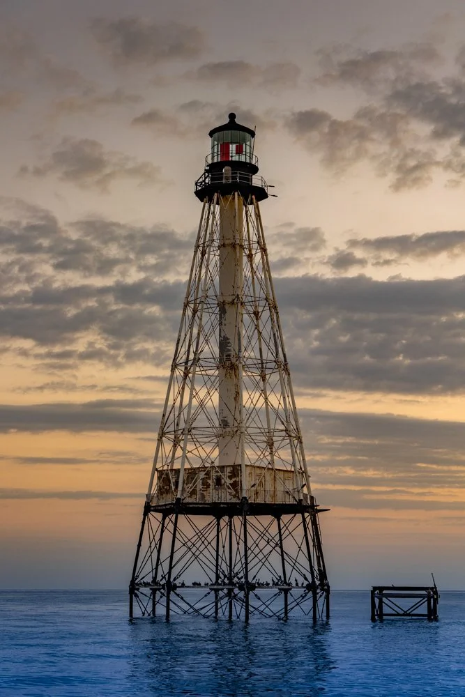 A tall, weathered lighthouse with a black top, standing in the water at sunset, surrounded by cloudy sky.