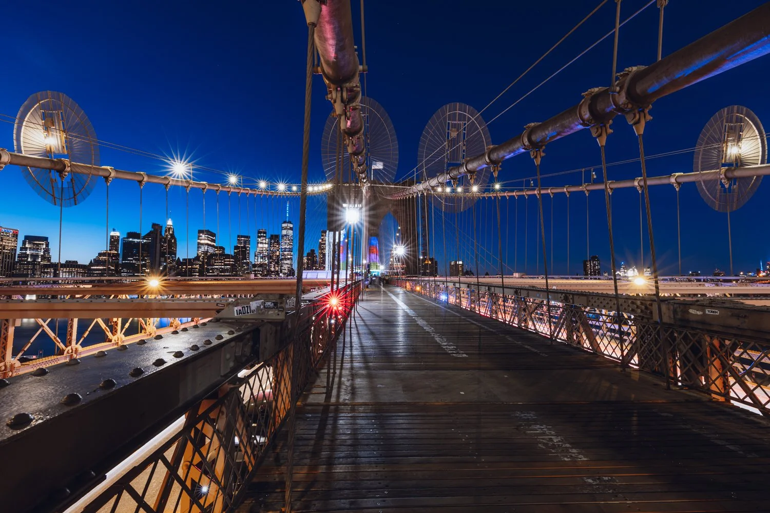 Night view of Brooklyn Bridge with Manhattan skyline in the background, showing illuminated buildings and the bridge's pedestrian walkway and cables.