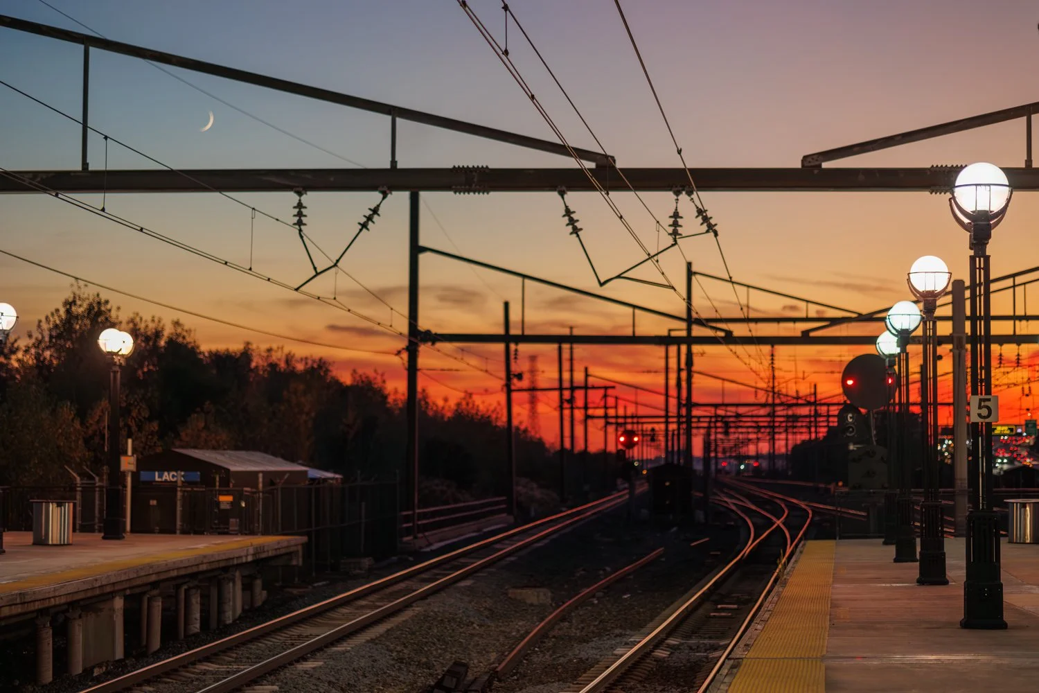Empty train platform at sunset with parallel train tracks, overhead electrical lines, and street lamps, and a crescent moon in the evening sky.