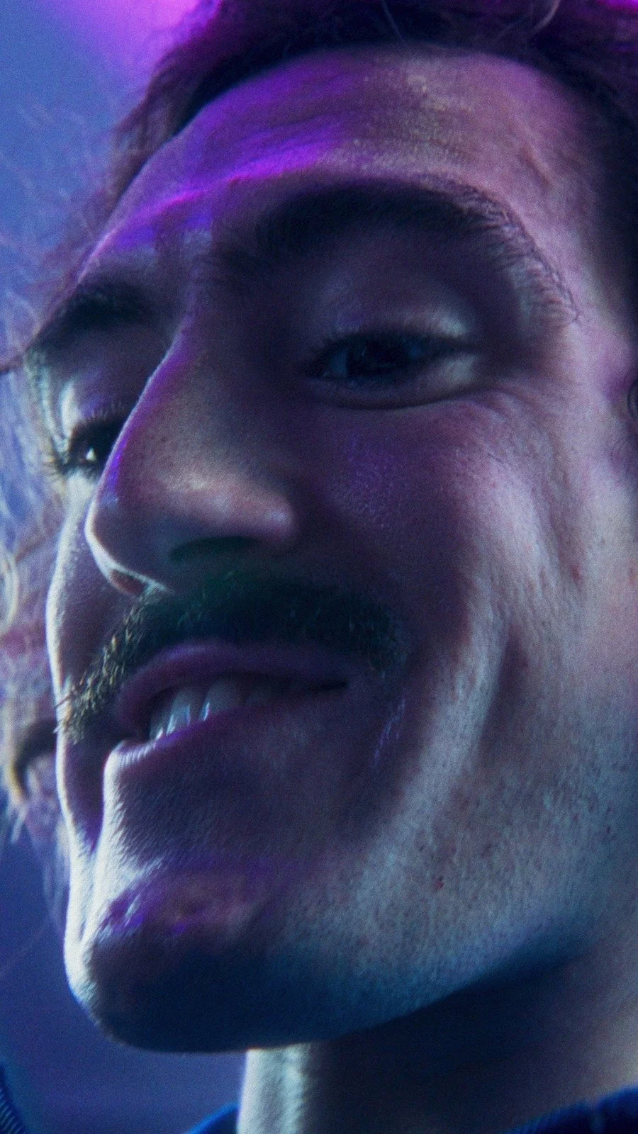Close-up of a man with a mustache smiling, illuminated by colorful purple and blue lighting.