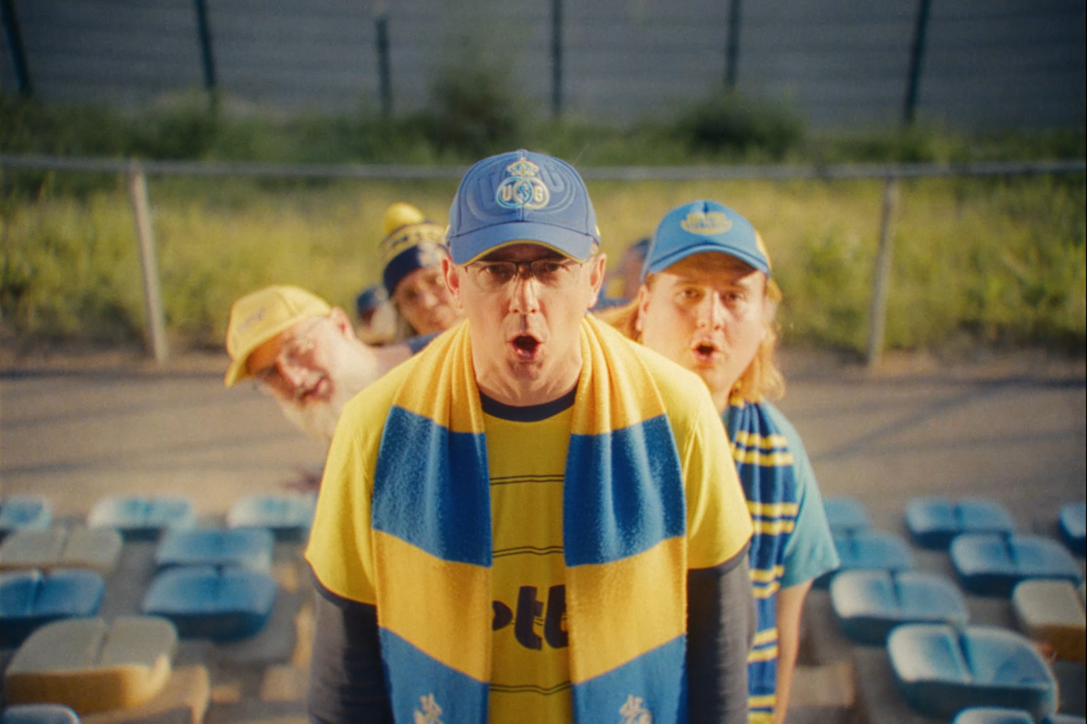 A group of men dressed in yellow and blue sports fans at an outdoor stadium, with stadium seats and green fencing in the background.