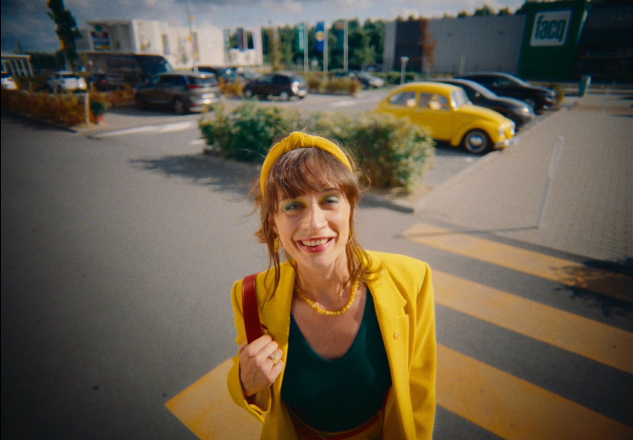 Smiling woman with red hair, wearing a yellow blazer, yellow headband, and green top, standing on a crosswalk in a parking lot, holding a red strap with her left hand.