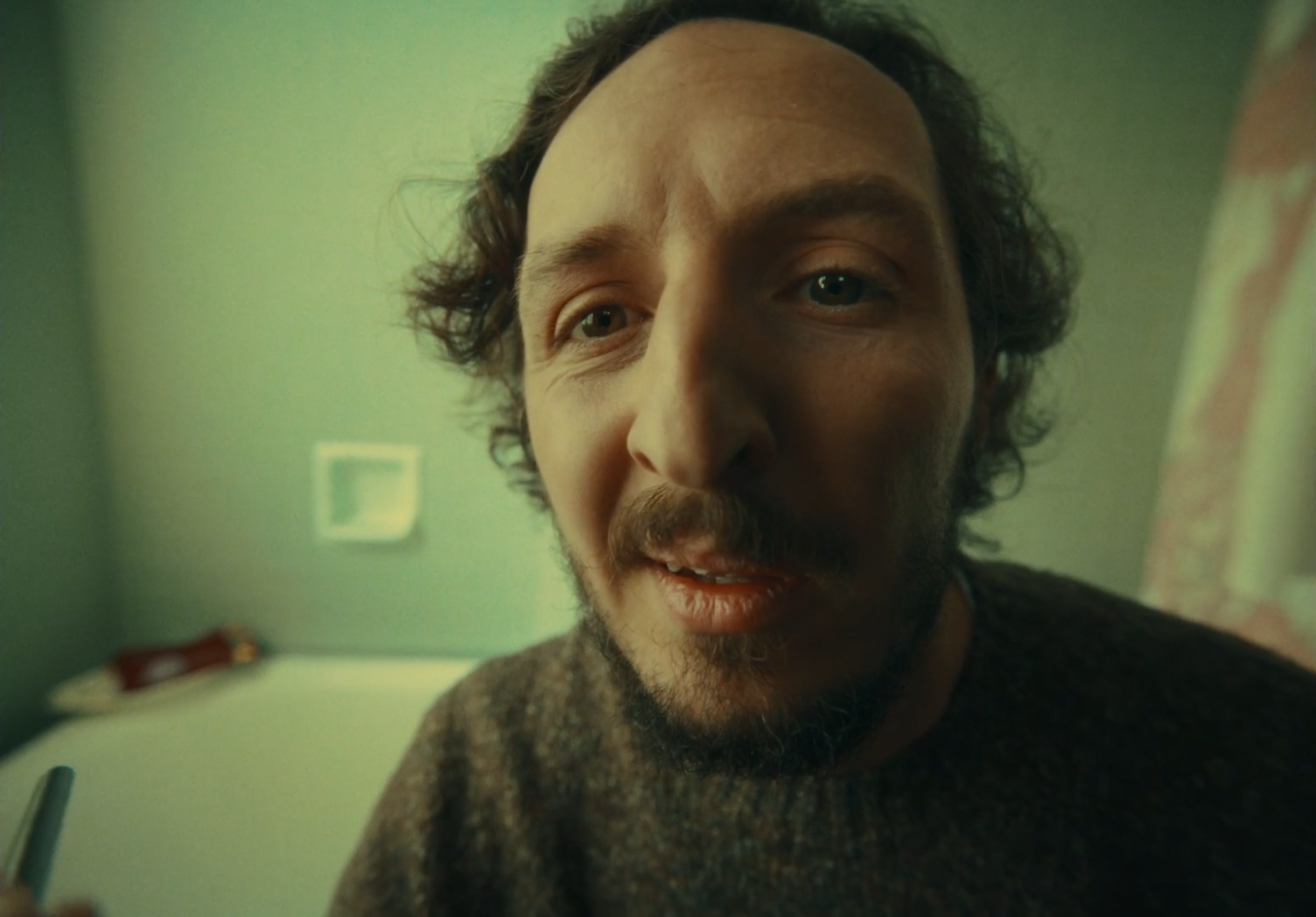 Close-up of a man with curly hair and a beard, looking into the camera, in a room with green walls and a bed in the background.