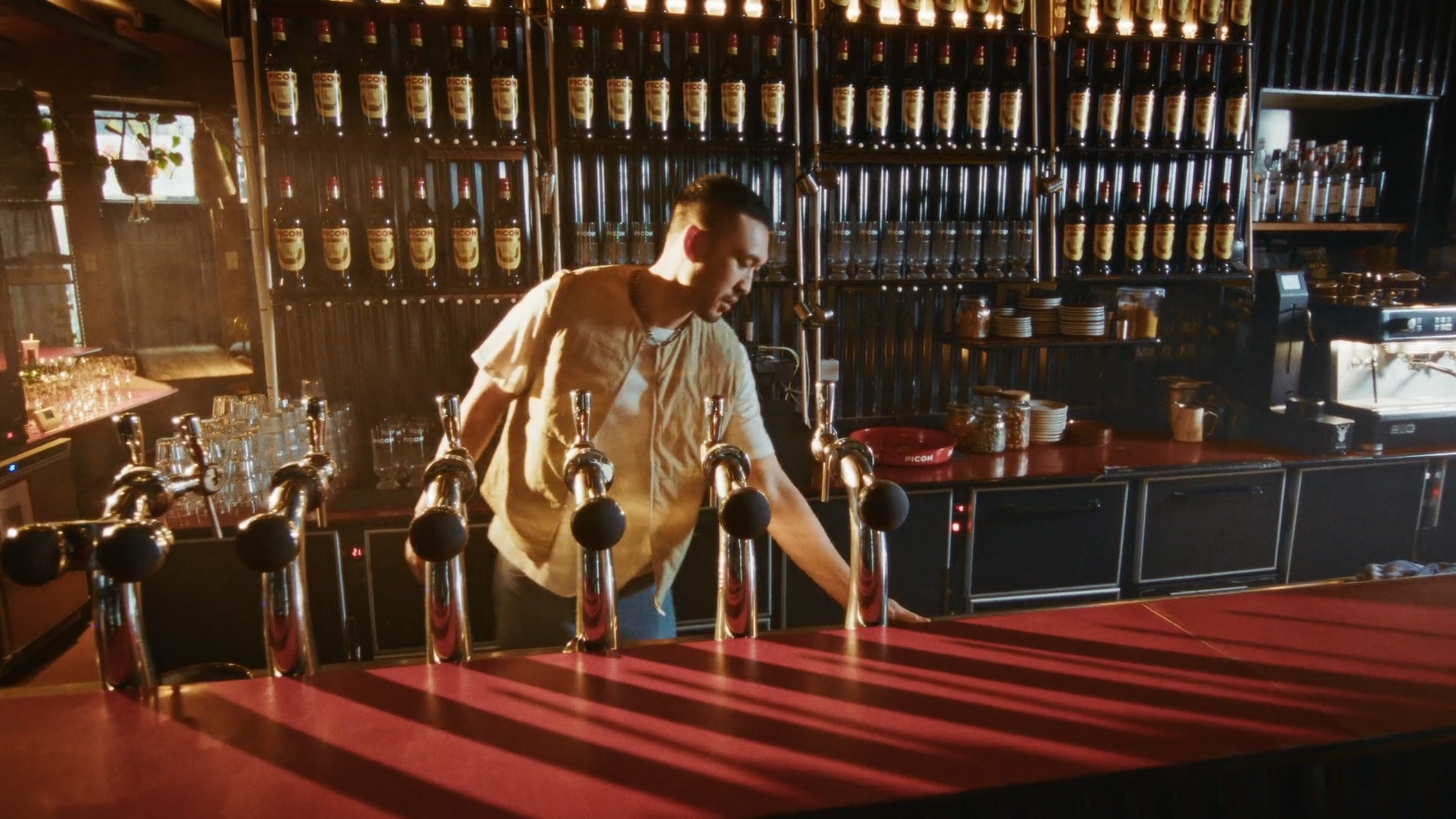 A bartender cleaning a bar counter with multiple beer taps and shelves of bottles in the background.
