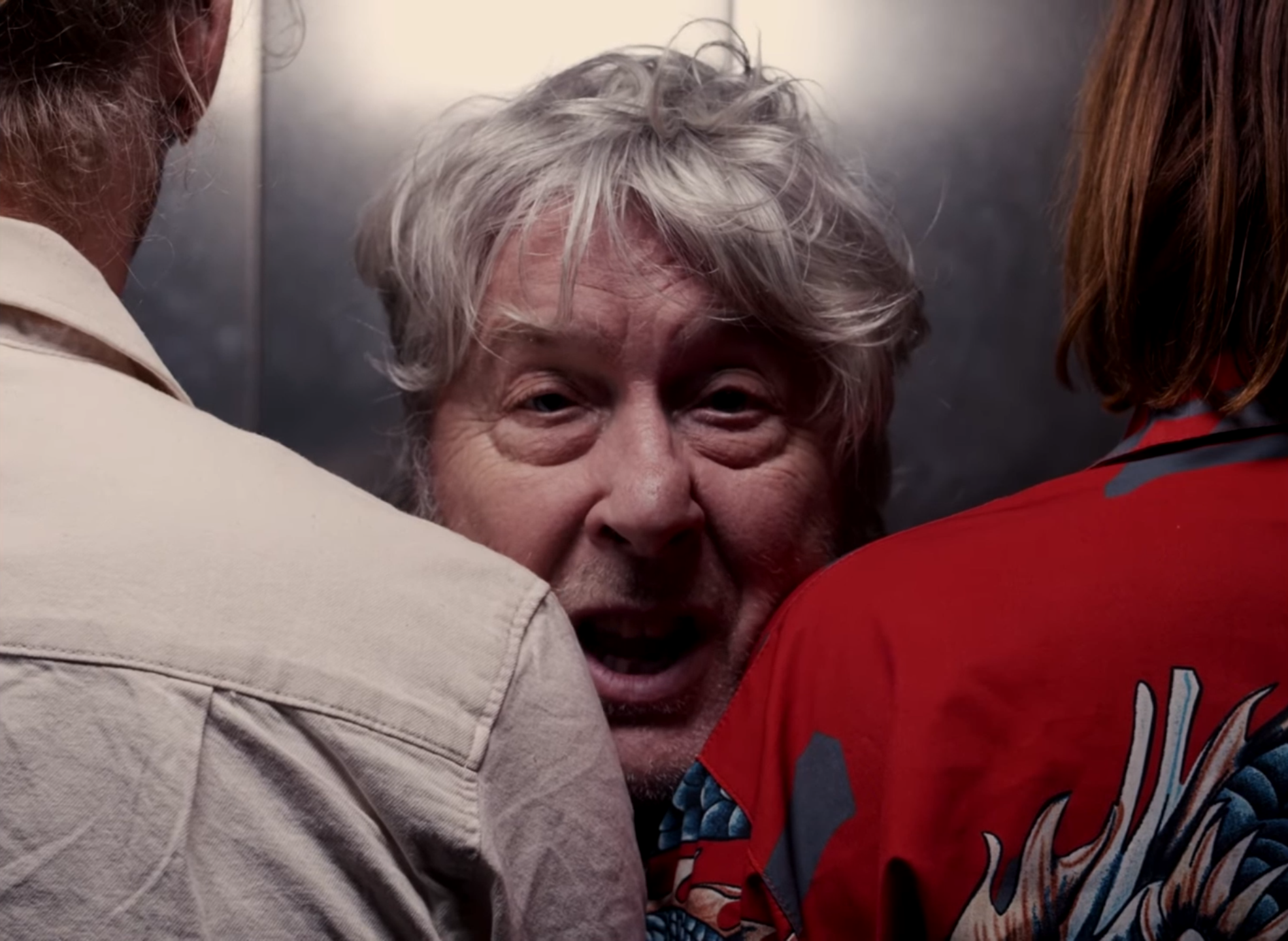 A man with gray hair is surrounded closely by two people, one on each side. The man appears to be in an elevator or a small enclosed space.