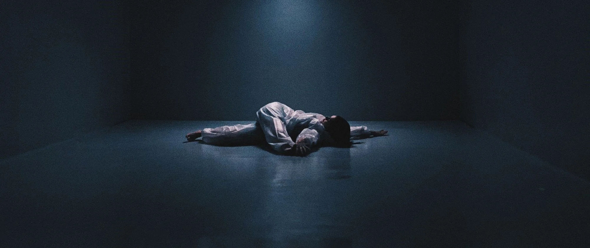 A person lying on the floor in a dark, empty room, curled up on his side with arms extended outward, illuminated by soft light from above.