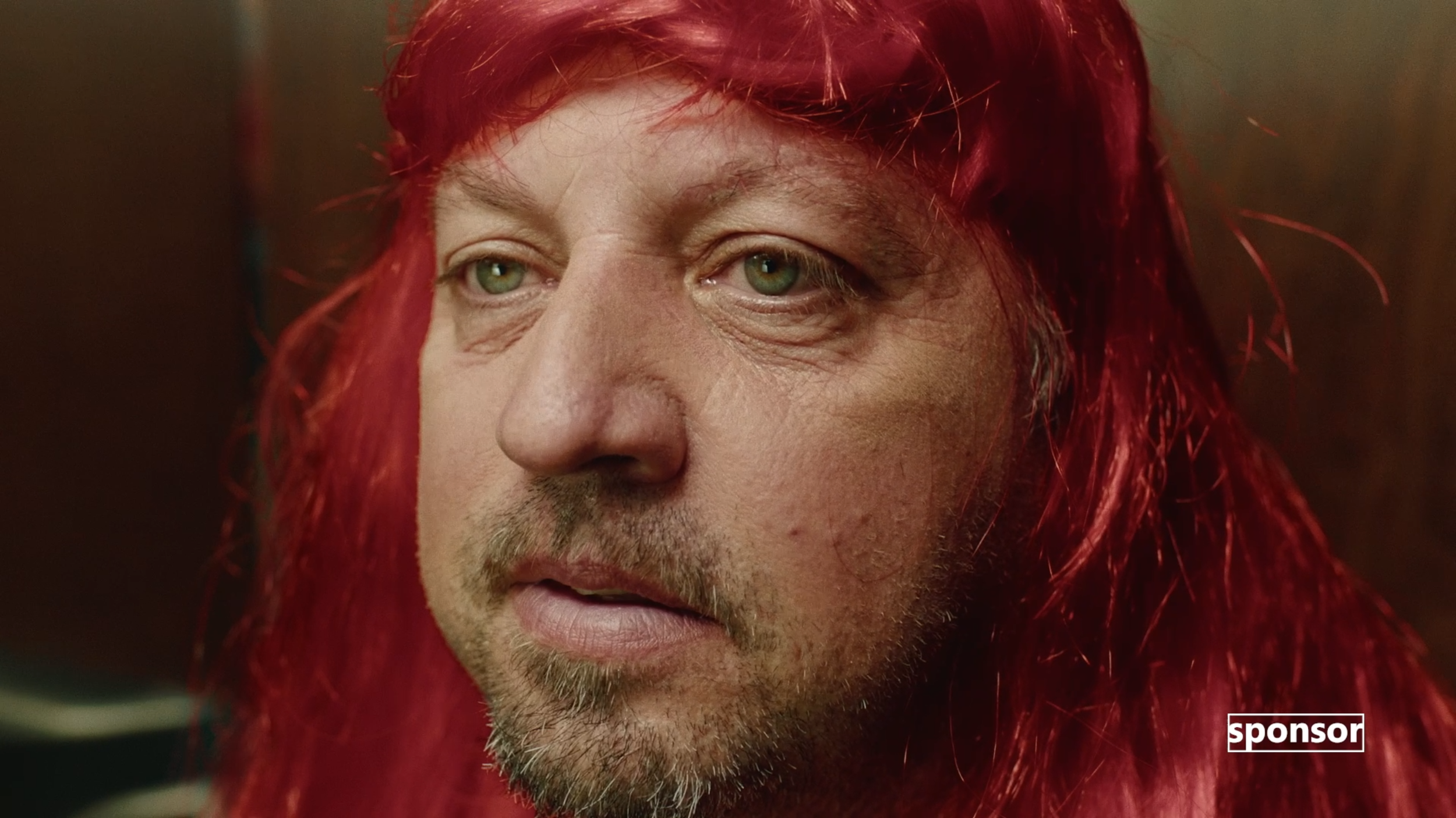 A close-up of a man's face with long red hair and green eyes, looking to the side with a serious expression.