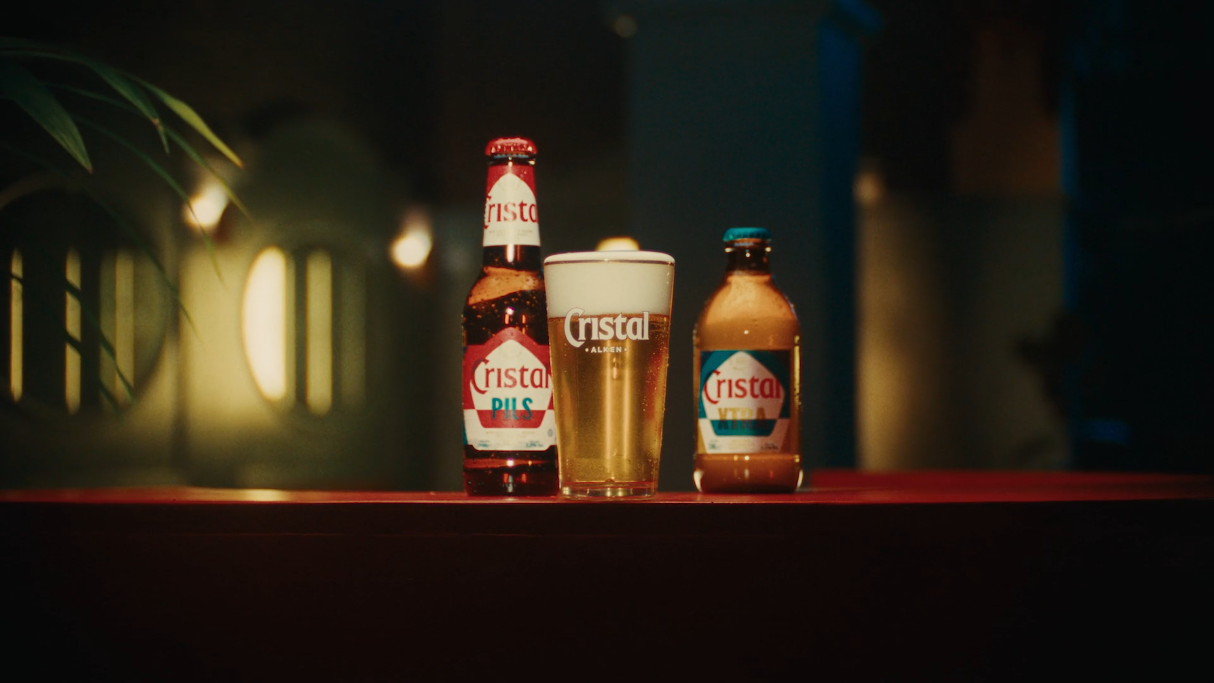 Two bottles of Cristal beer and a glass of beer on a red table, with a blurred background of string lights and foliage.