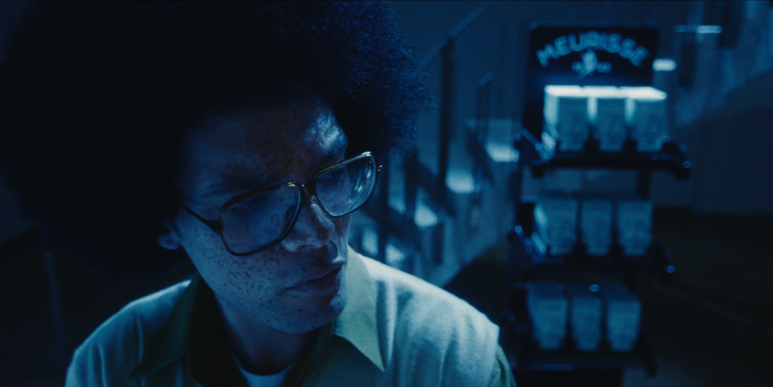 A person with glasses and freckles looking down in a dark, blue-lit environment with a roller cart in the background labeled 'MEURERS'.