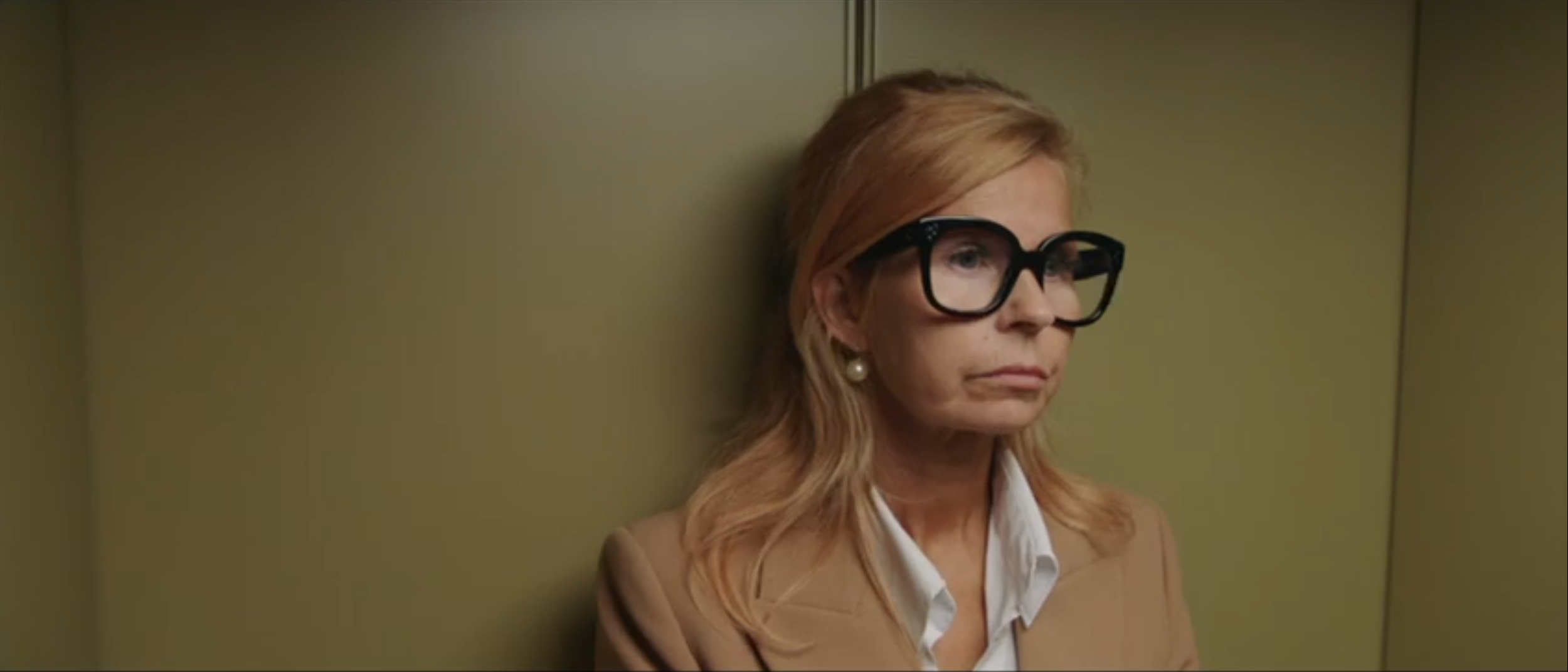 A woman with blonde hair wearing large black glasses, pearl earrings, a white shirt, and a tan blazer, standing inside an elevator with a green wall.