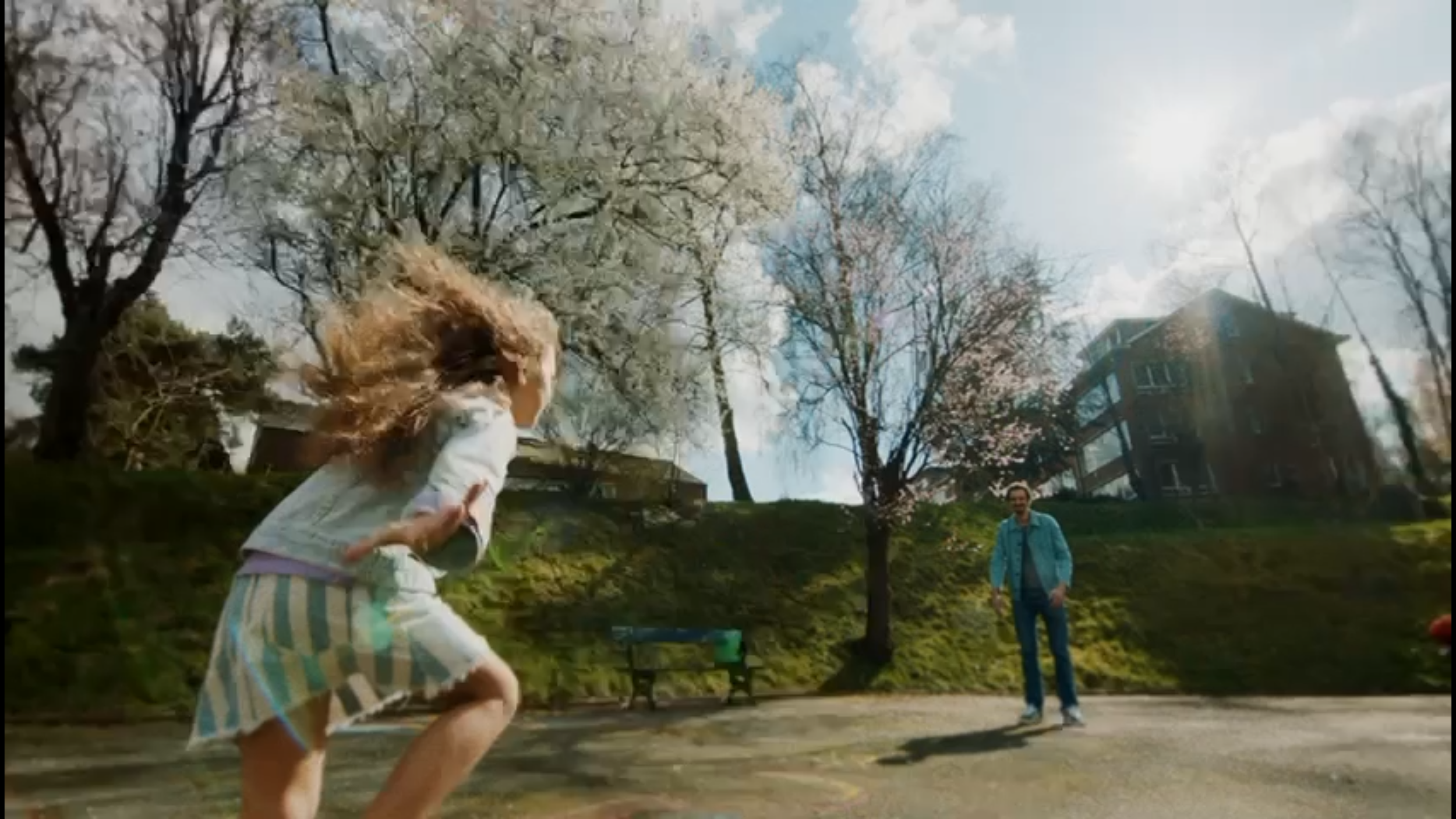 A girl with curly hair running or jumping on a park pavement with a man standing in the background. Trees with blossoms are visible, and a building is seen in the distance with a bright sun shining.