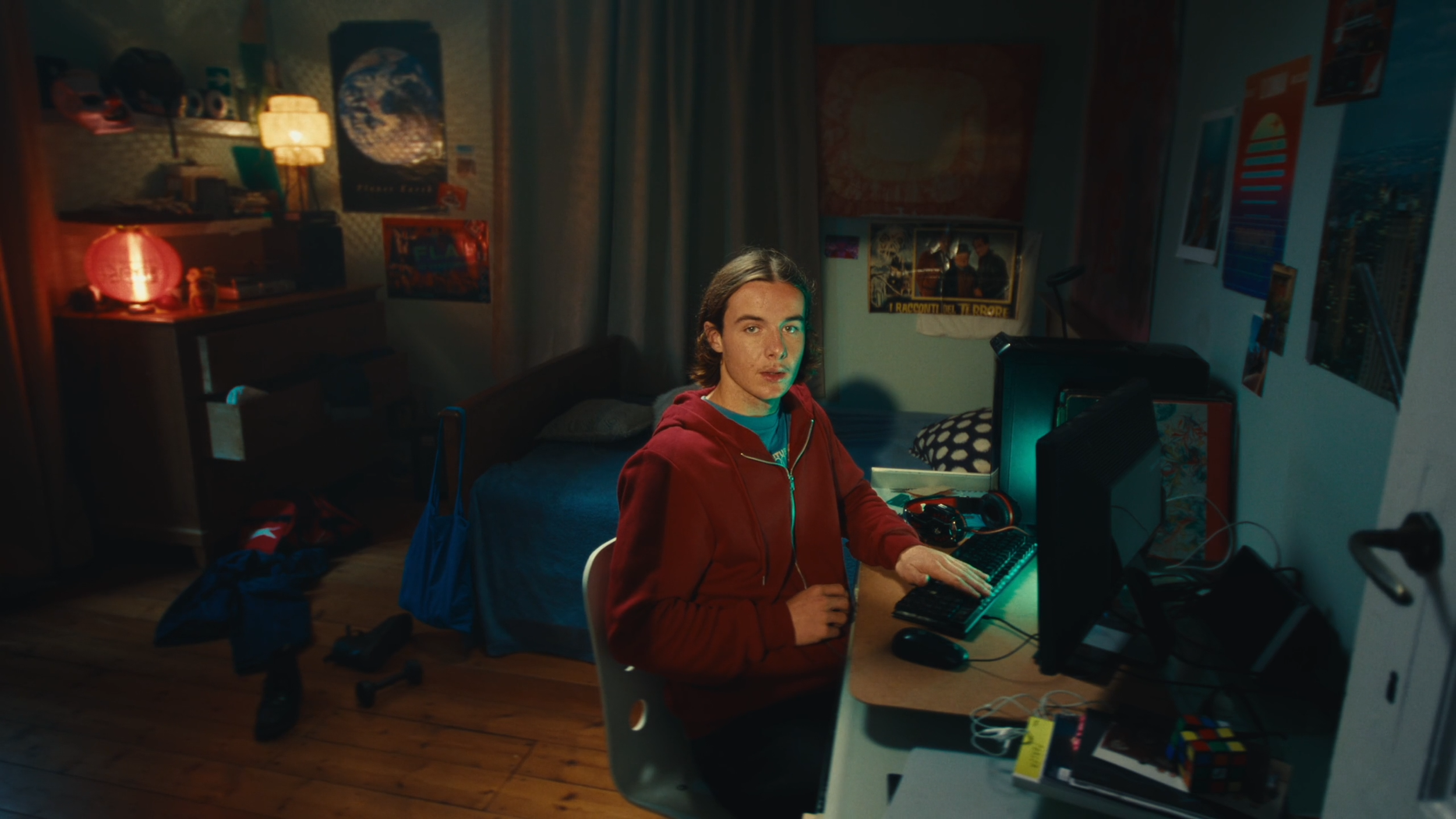 A young man with shoulder-length hair, wearing a red hoodie, sitting at a desk with a computer in a dimly lit bedroom, surrounded by posters and various personal items.