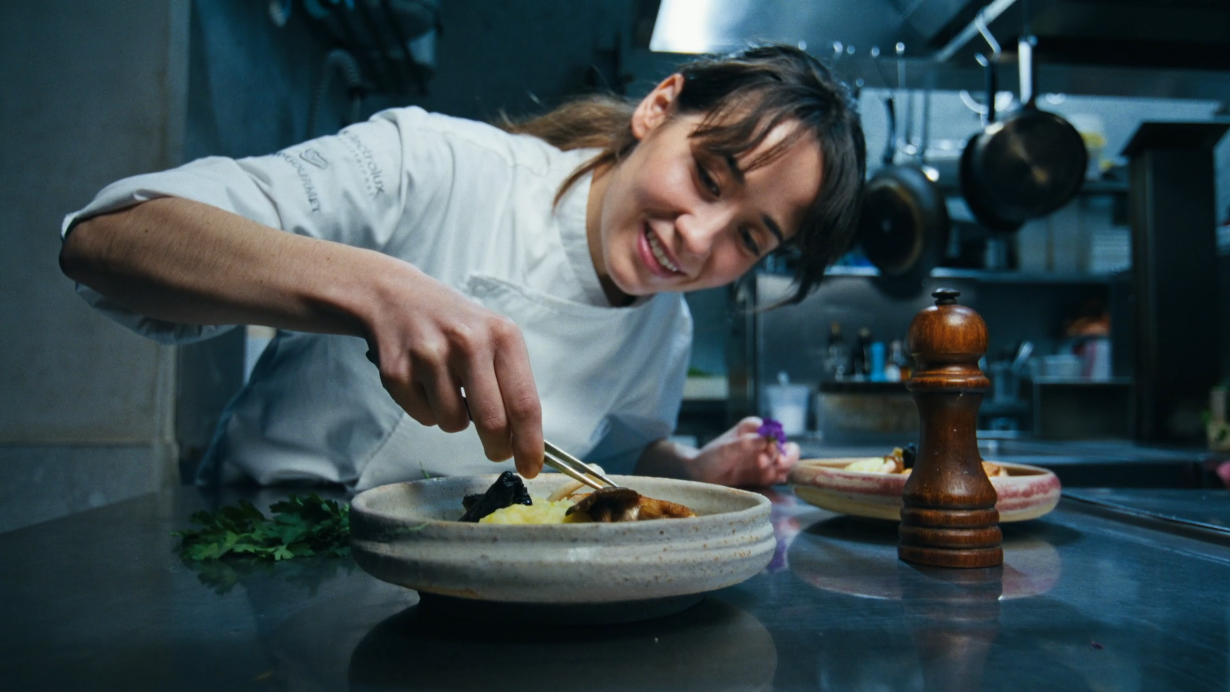 A female chef in a white uniform carefully garnishes a dish with tongs in a professional kitchen.