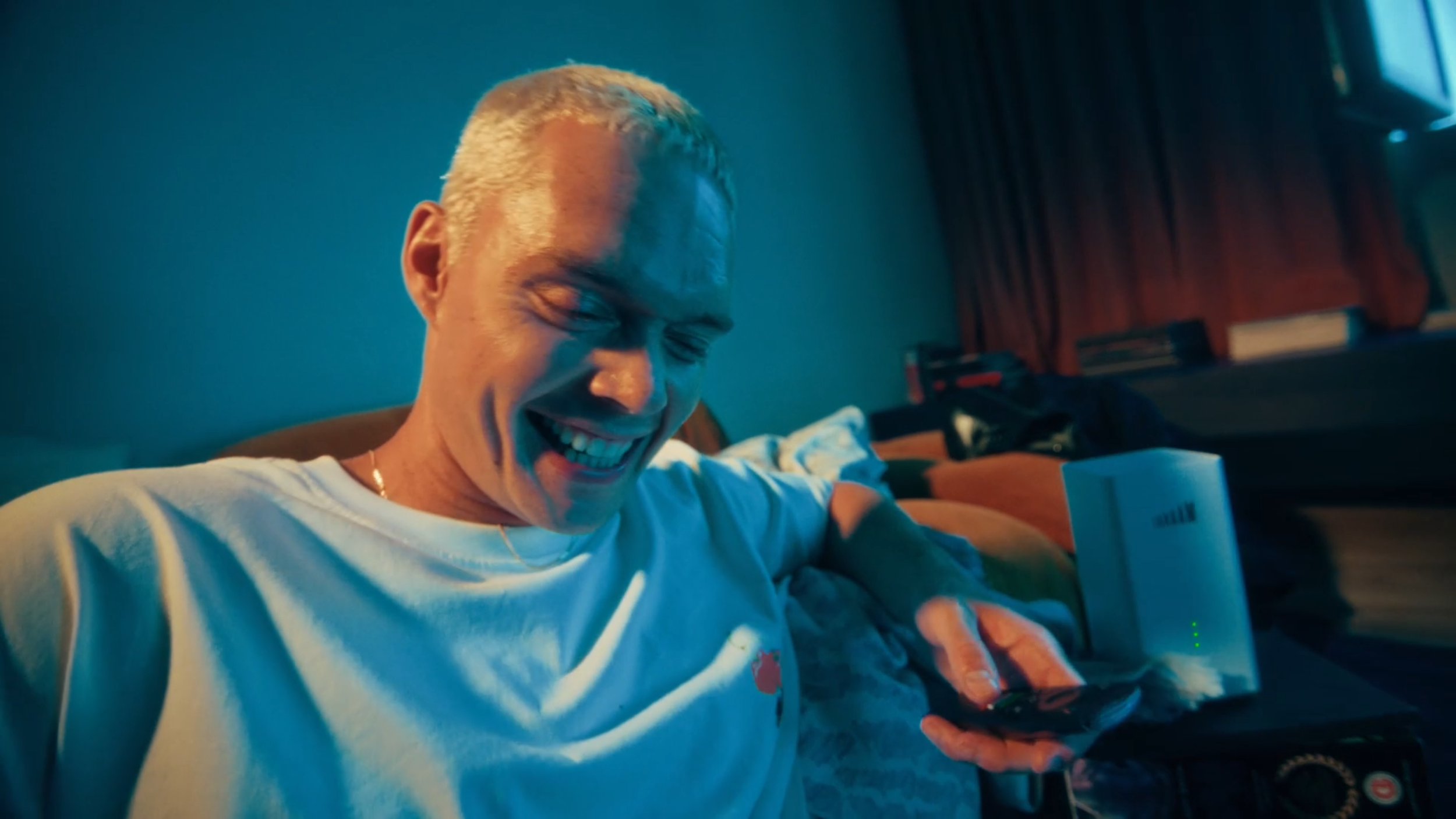 A young man with blond hair smiling while looking at his phone in a dimly lit room with blue tones, sitting on a couch with a orange cushion, with a white Wi-Fi router and a stack of books in the background.