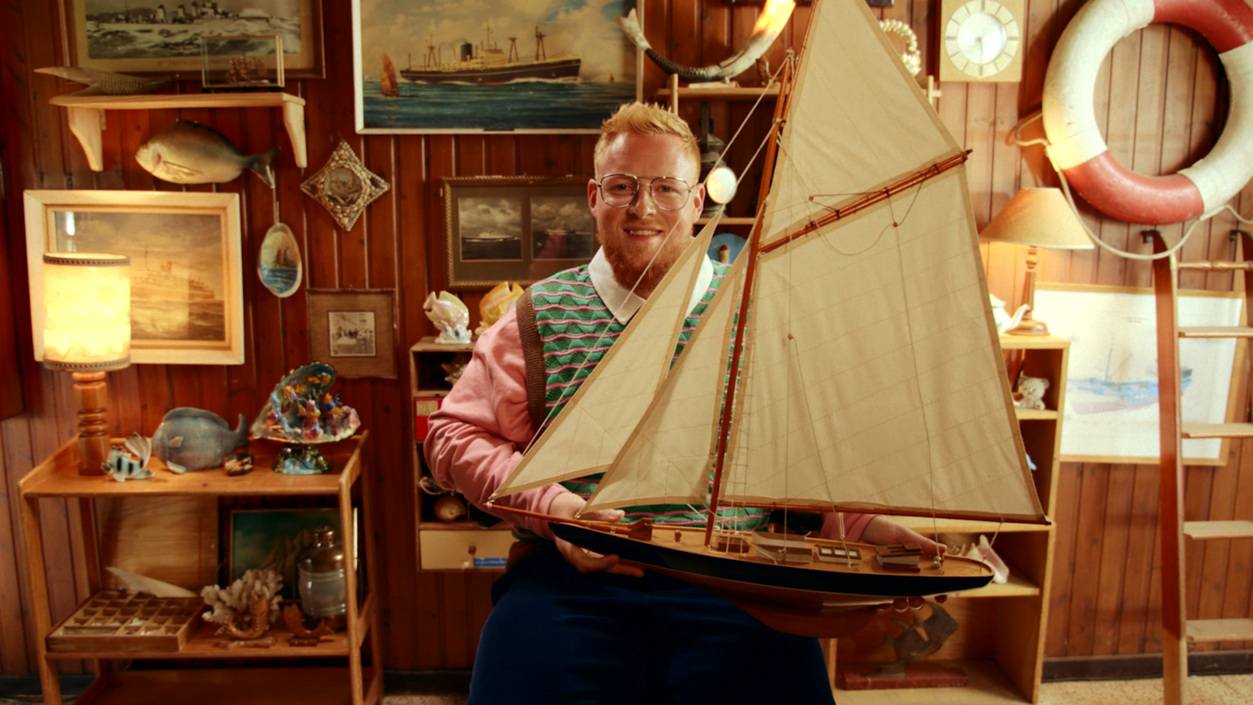 A man with glasses, wearing a colorful sweater vest, holding a large model sailboat indoors with wooden walls decorated with nautical-themed pictures and objects, including a fish, a life ring, and various maritime decorations.