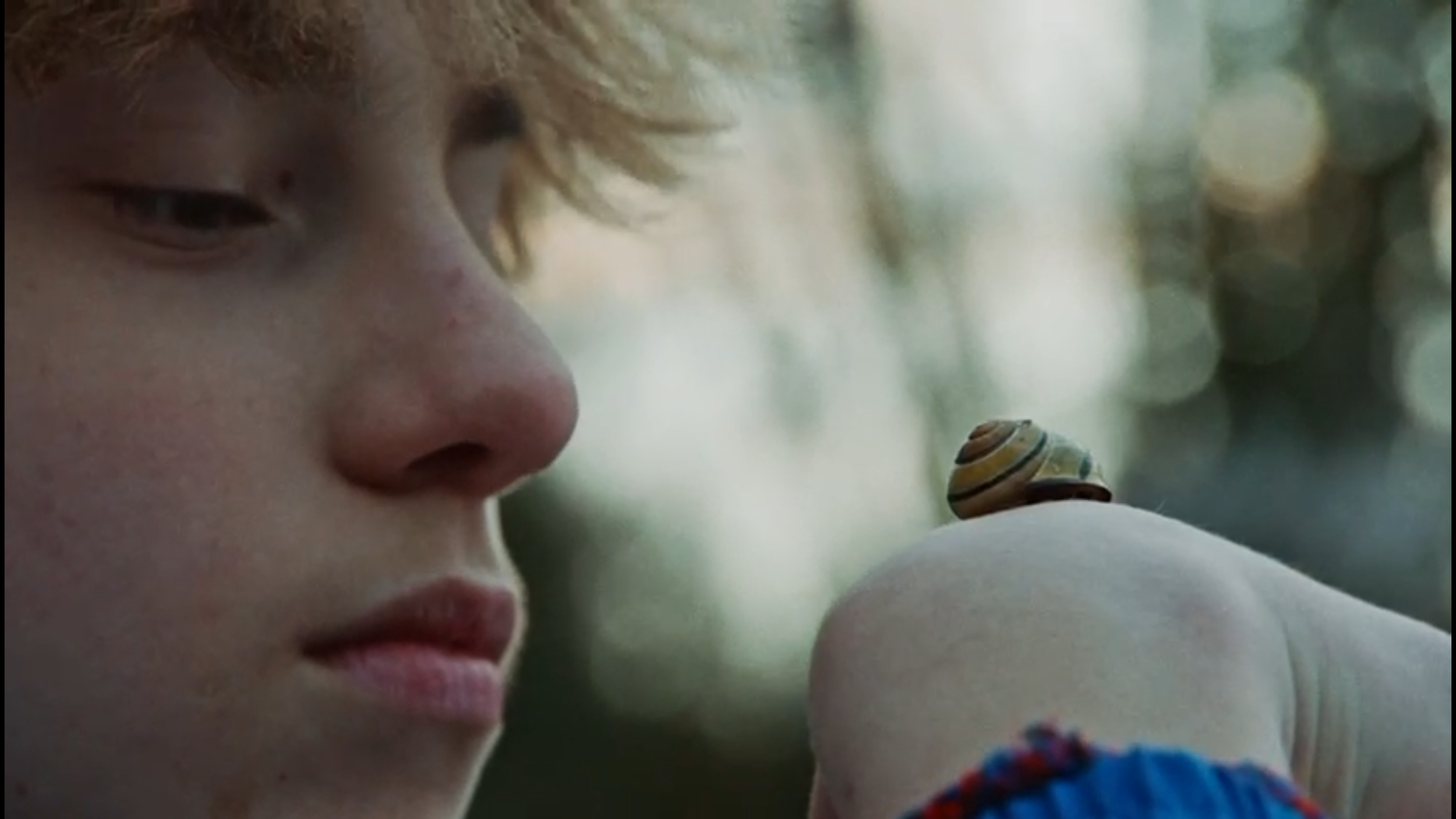 Close-up of a young boy with light skin and blonde hair, looking intently at a small snail on his arm, with a blurred outdoor background.