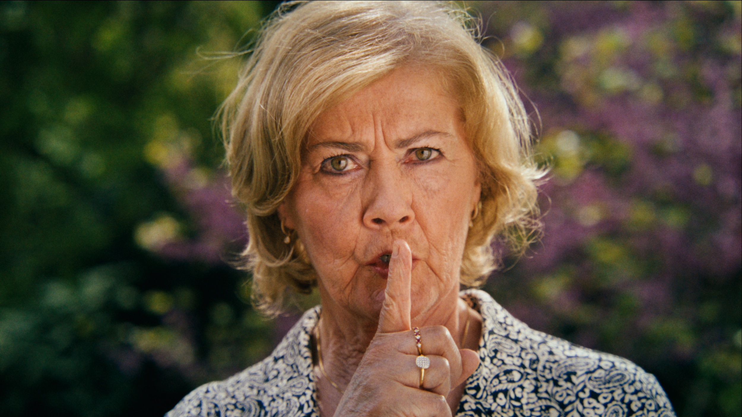 An elderly woman with blonde hair making a shushing gesture with her finger on her lips, outdoors with a blurred background of trees.