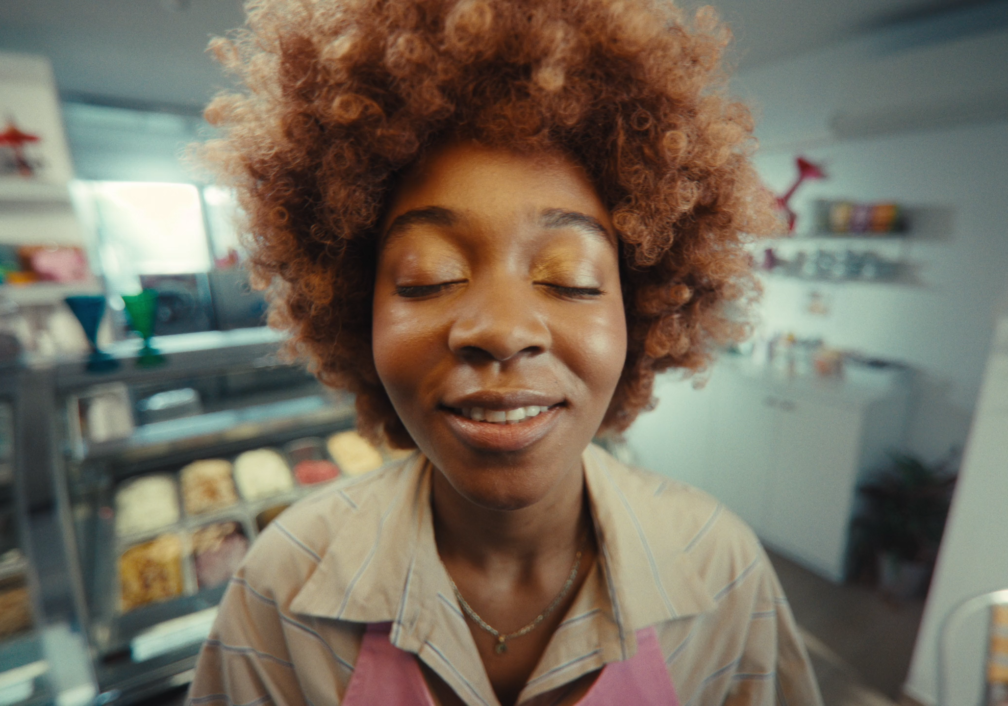 A woman with curly reddish-brown hair smiling with closed eyes in a bakery or cafe setting.