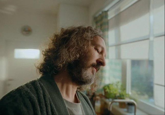 A man with curly hair and a beard looking downwards in a brightly lit room near a window.