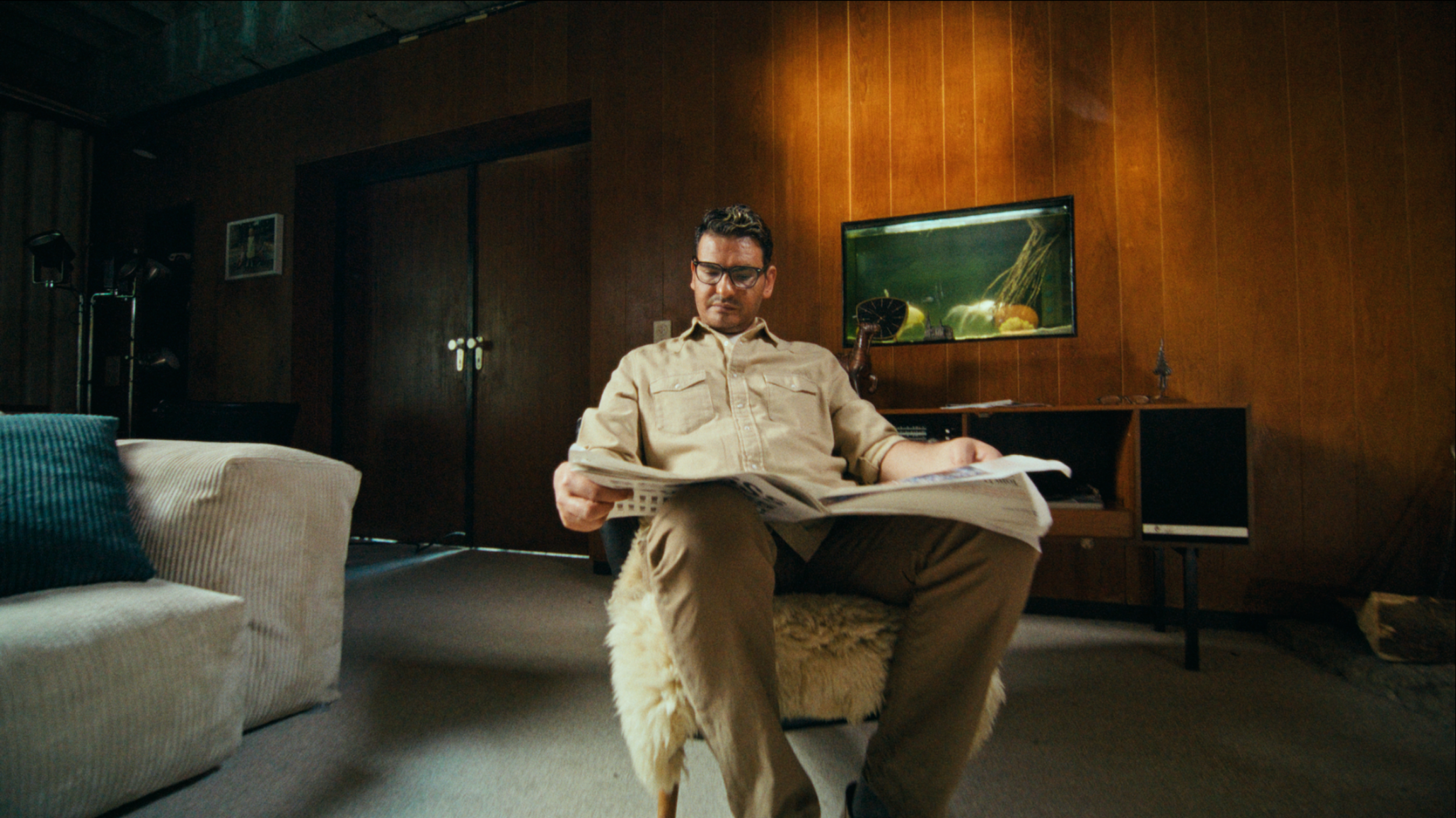 Man wearing glasses and a beige shirt sitting on a furry chair, reading a newspaper in a room with wood-paneled walls, a sofa with a blue pillow, and a TV in the background.