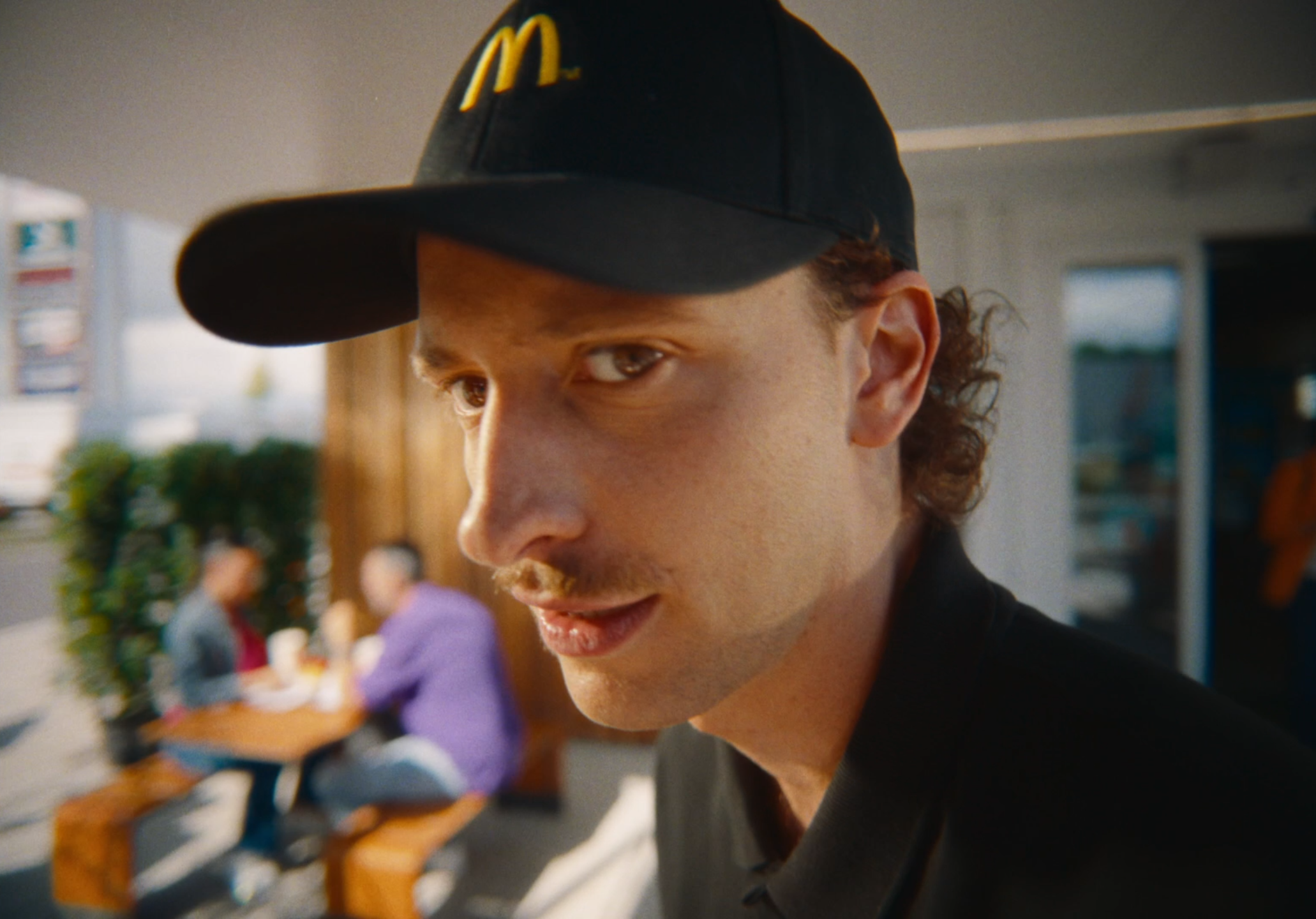 Close-up of a man wearing a black cap with an 'McDonald's' logo, looking at the camera with a slight smile, outdoors with people sitting at a table in the background.