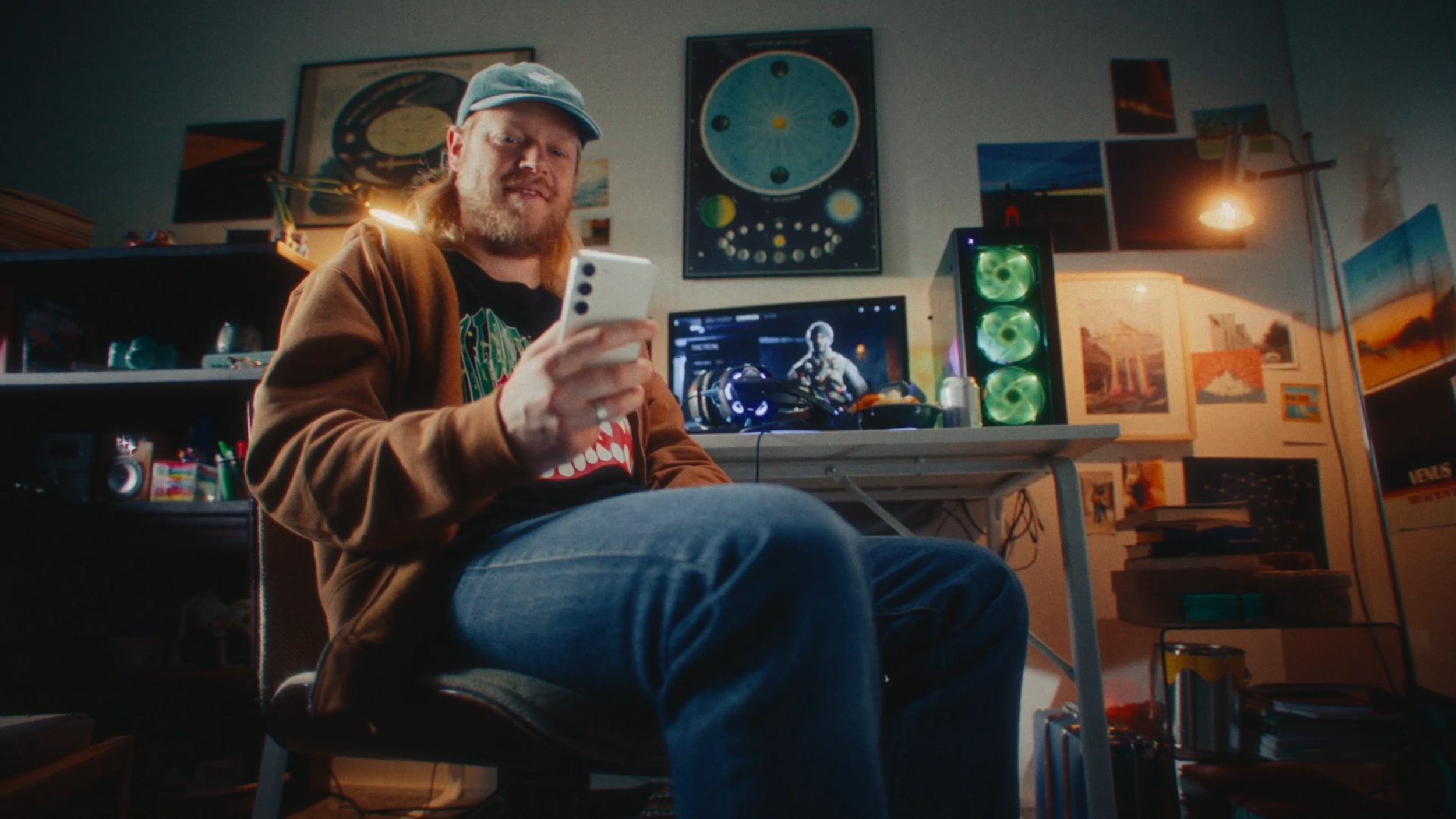 A young man with long hair and a beard sitting in a cluttered room, looking at his smartphone. Behind him, a computer monitor displays a sci-fi character and a gaming setup with green lights. The room has various posters and pictures on the walls, an