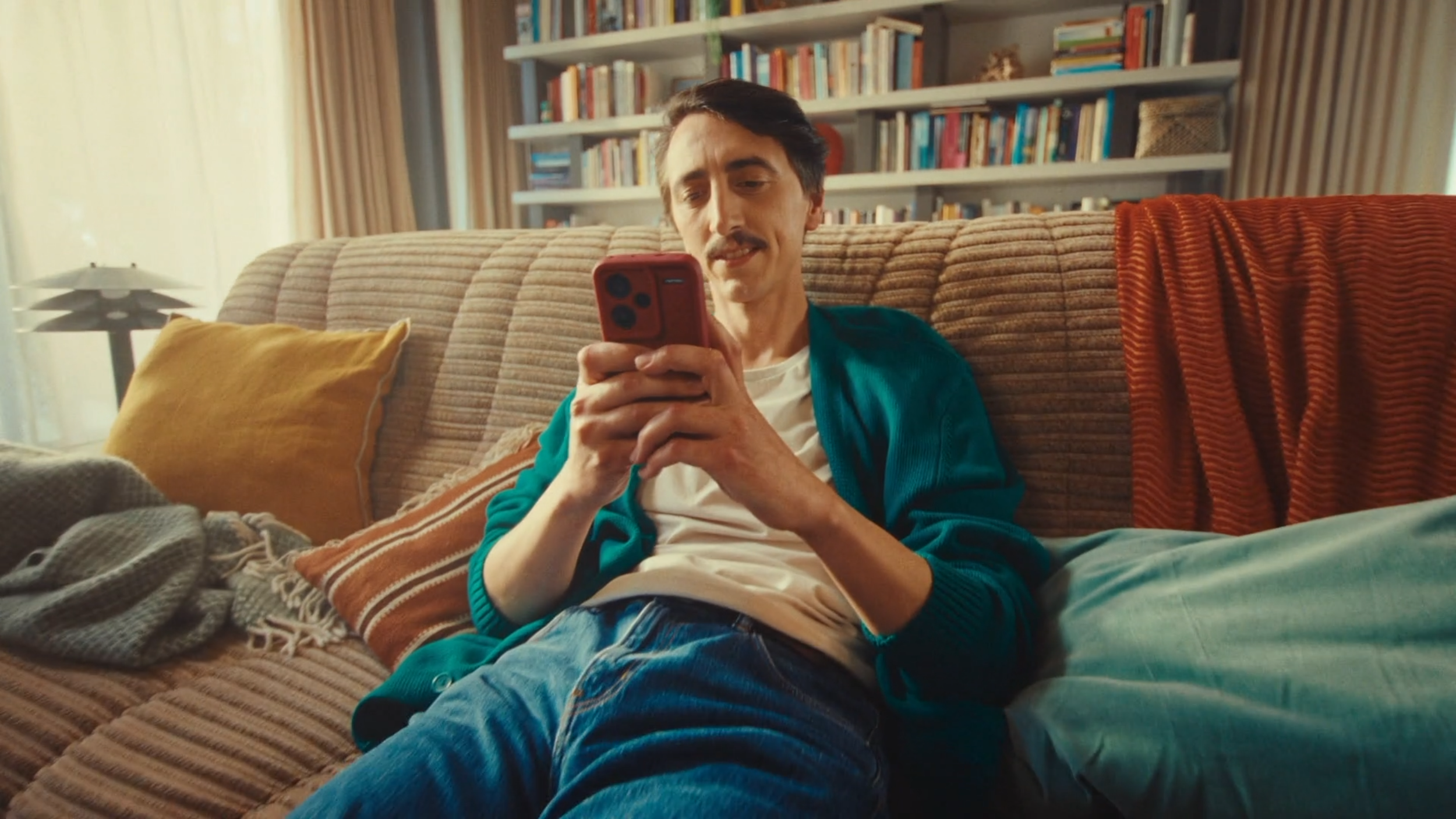 A man with dark hair, mustache, and light skin sitting on a beige sofa, looking at his red smartphone in a living room with a bookshelf in the background. There are yellow and orange pillows on the sofa, and a striped blanket on the left side. Sunlig